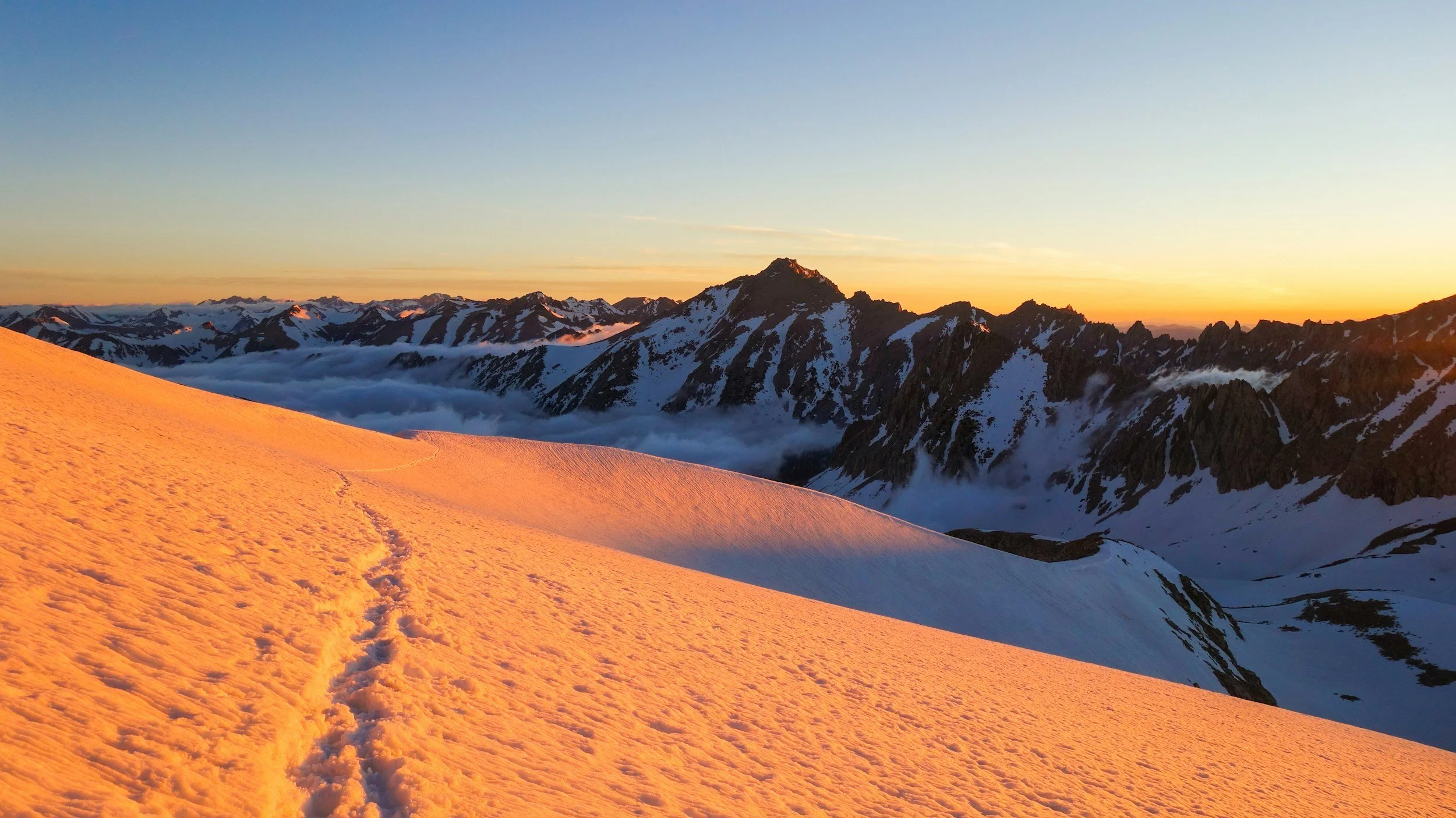 Snow-covered mountain slope with footprints in the foreground, jagged mountain peaks in the distance, and a sunset sky with colors ranging from orange to blue.
