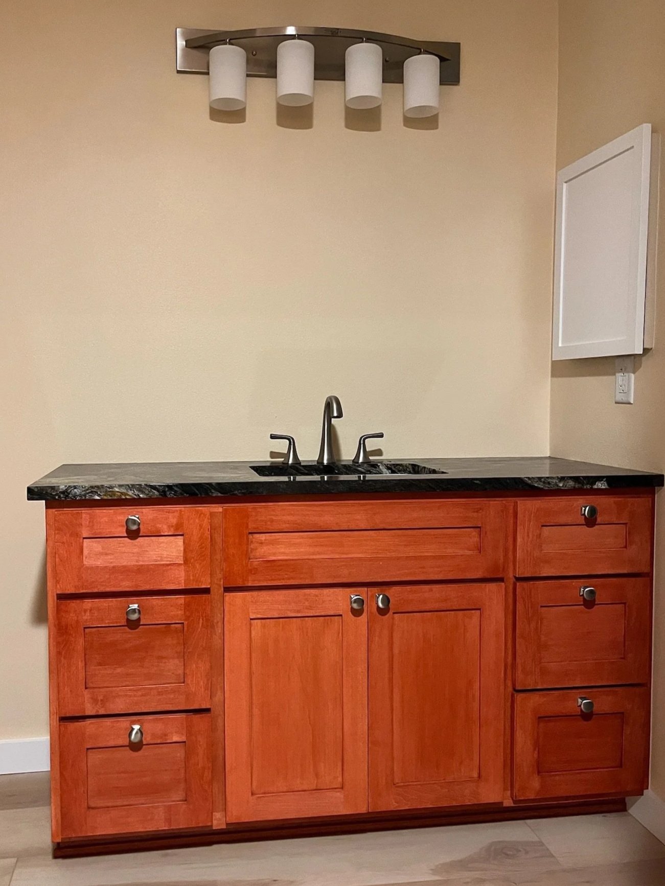 A kitchen cabinet with a dark granite countertop, a black sink, and a brushed nickel faucet. The cabinet is made of warm reddish-toned wood with multiple drawers and doors. There is a wall-mounted white electrical panel on the right side and a modern ceiling light fixture with five cylindrical white shades above.