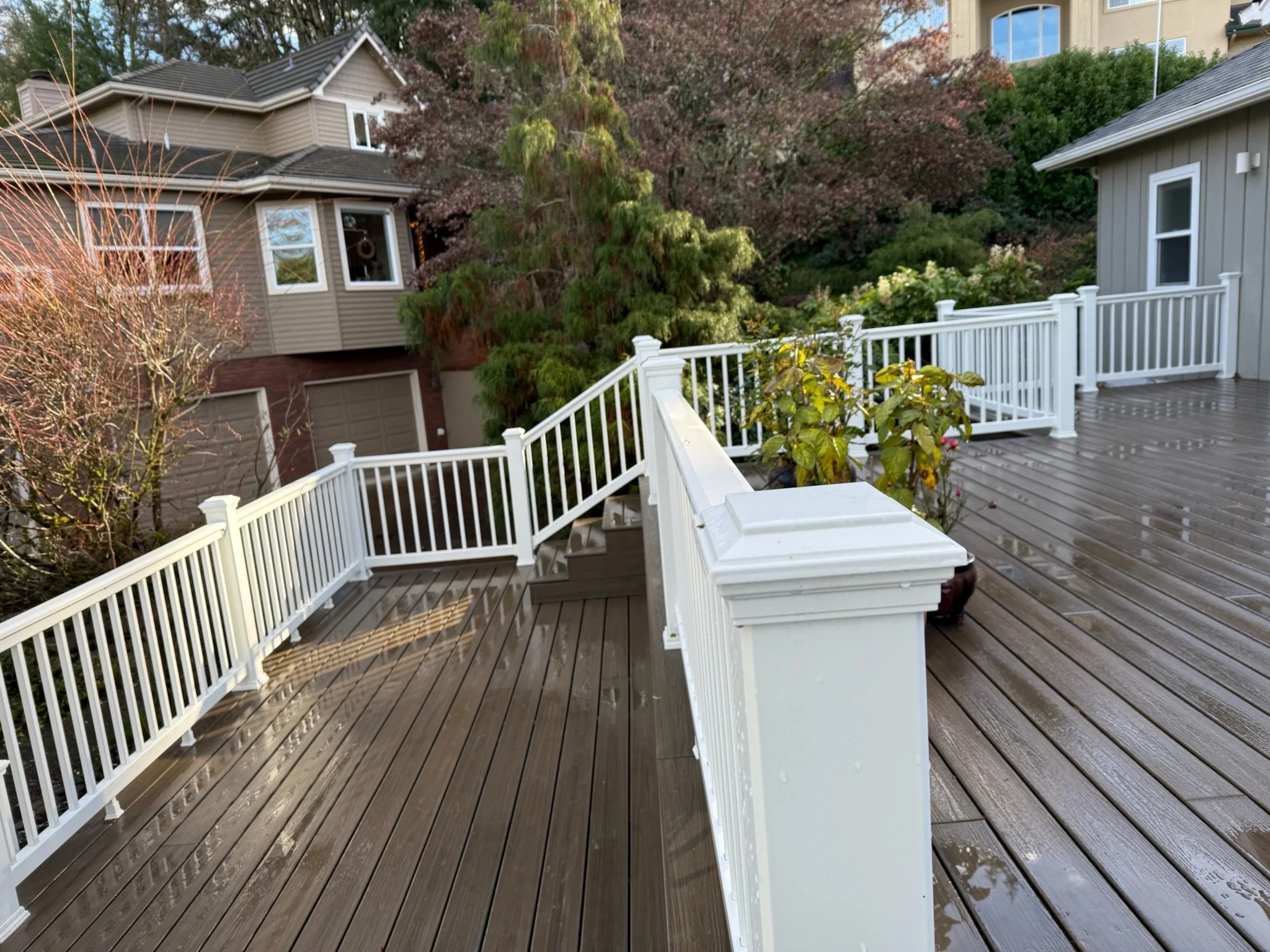 A wooden deck with a white railing overlooking a garden with trees and bushes, and a neighboring house in the background.