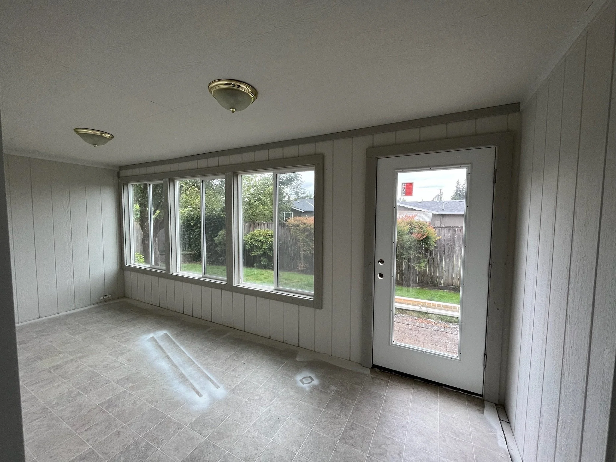 A sunroom with large windows and a door, white wooden panel walls, tiled flooring, and ceiling lights, with a view of a backyard with trees and a fence.
