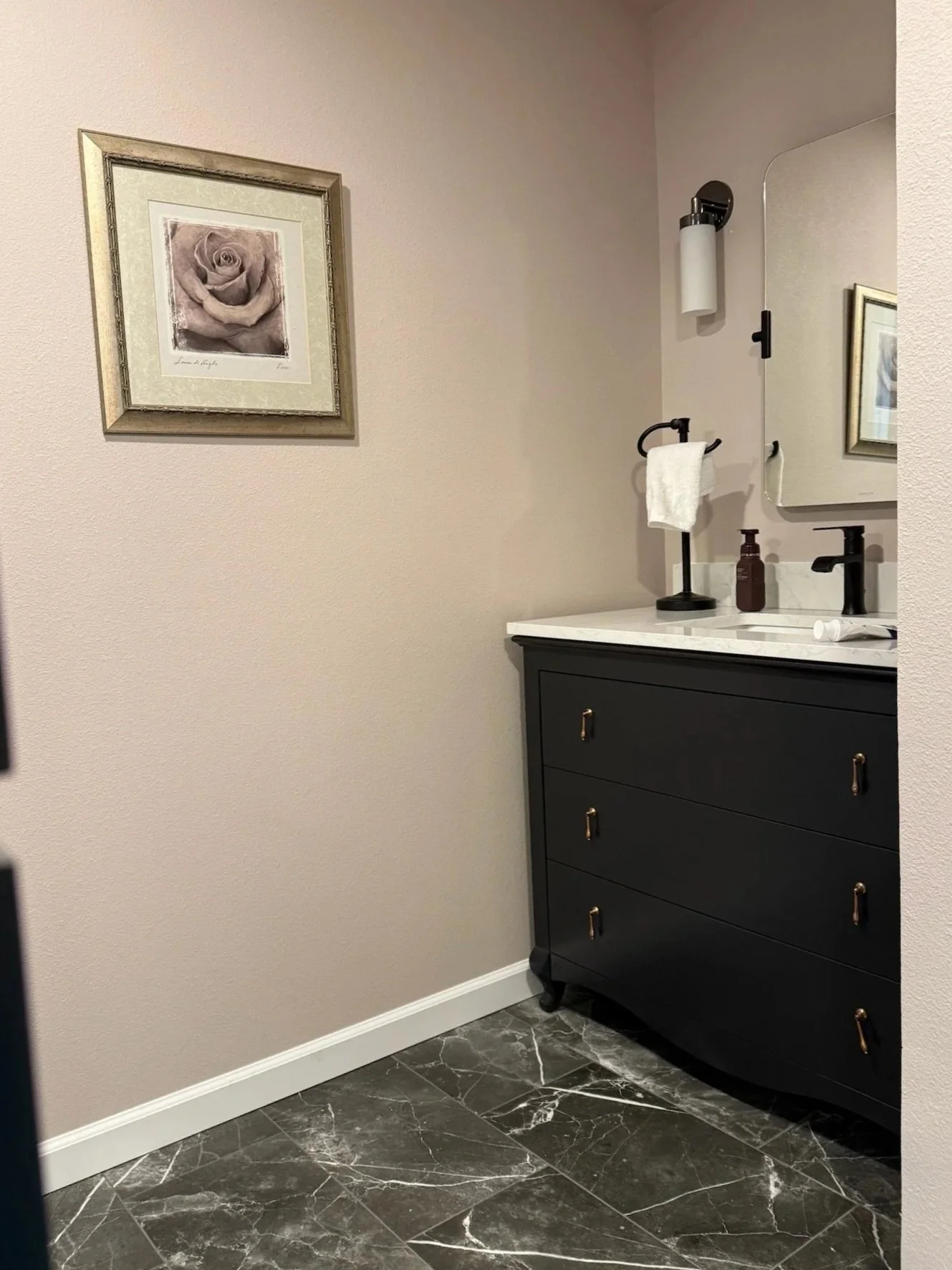 The image shows a bathroom corner with a black vanity cabinet, a white marble countertop, a large mirror, a wall-mounted light fixture, a soap dispenser, a pill bottle, a towel ring with a white towel, and a framed black-and-white photo of a rose on the beige wall. The floor is dark marble with white veining.