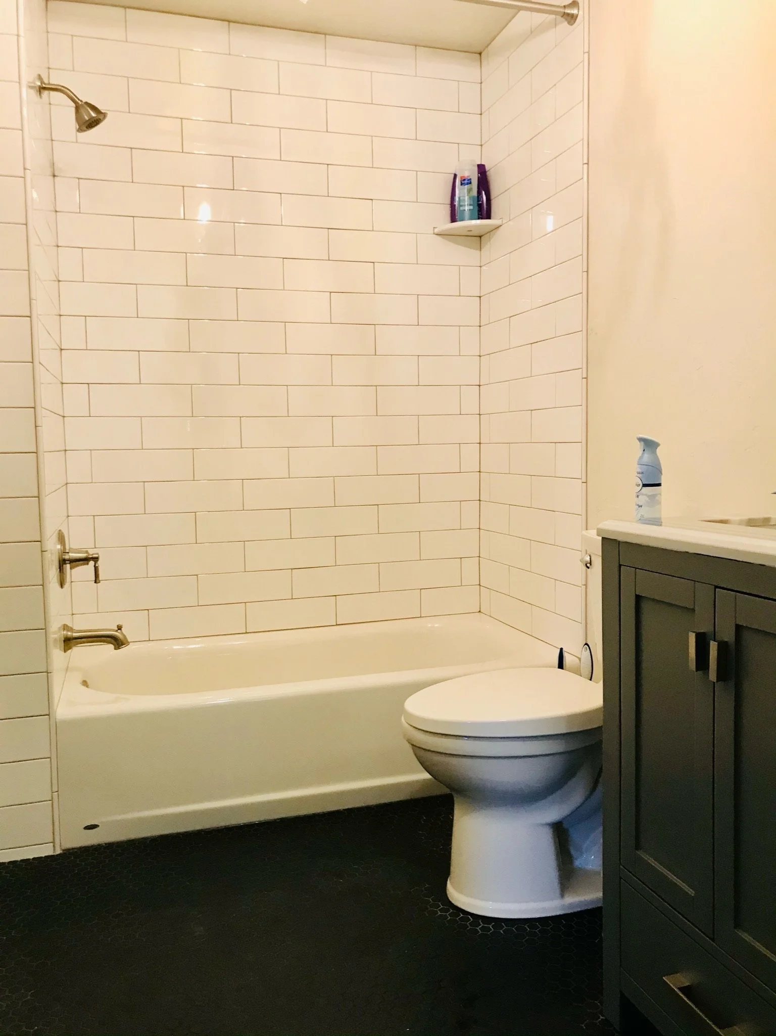 A bathroom with white subway tile walls, a bathtub, a toilet, a gray cabinet, and a black hexagon tile floor. There is a shelf with shampoo bottles and a spray bottle on the side of the bathtub.