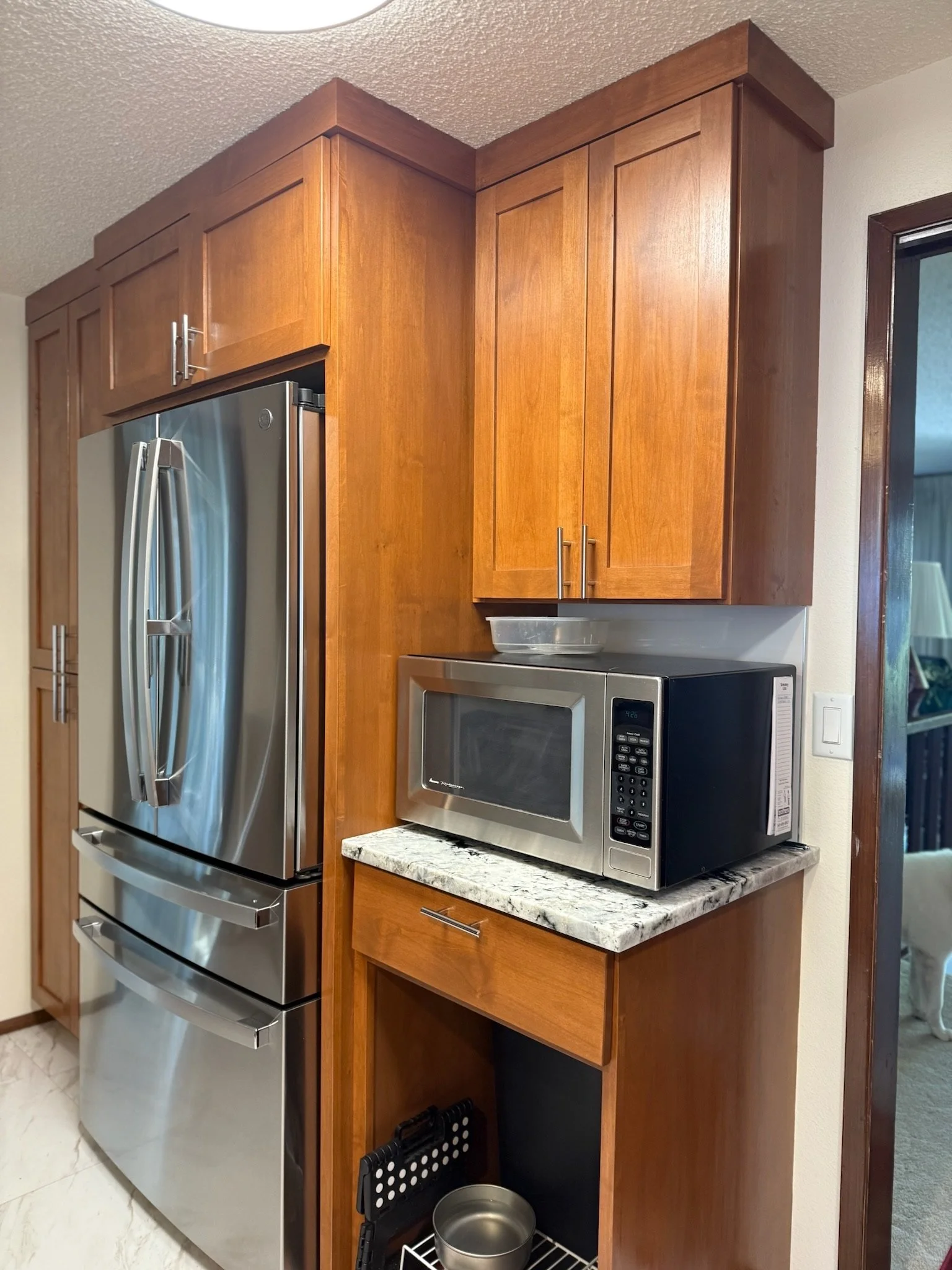 Kitchen corner with wooden cabinets, stainless steel refrigerator, microwave oven on granite countertop, and dog food bowl on a rack.