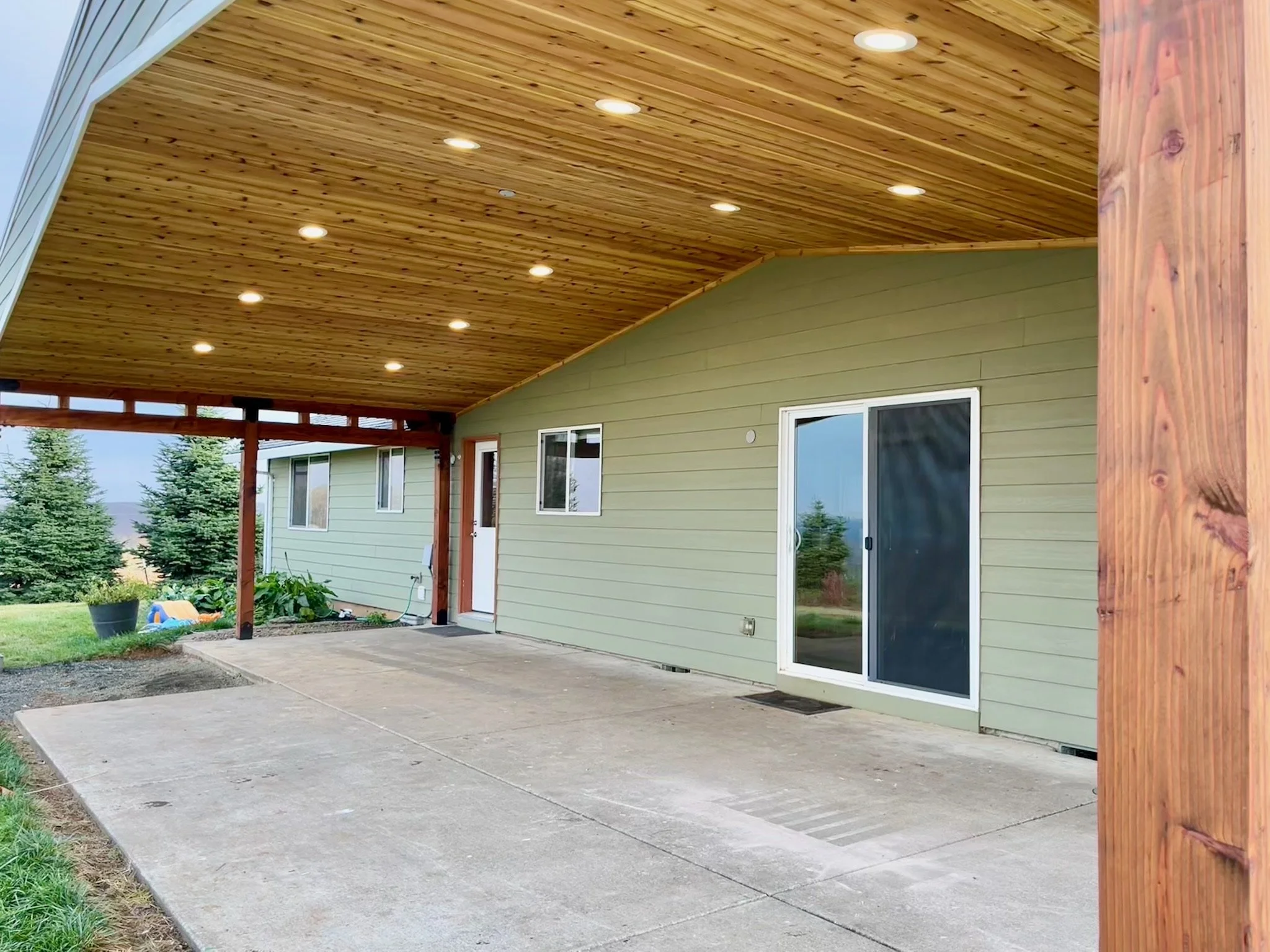 Backyard patio area with concrete flooring, a green house with sliding glass doors and windows, and a wooden ceiling with recessed lighting, surrounded by trees.