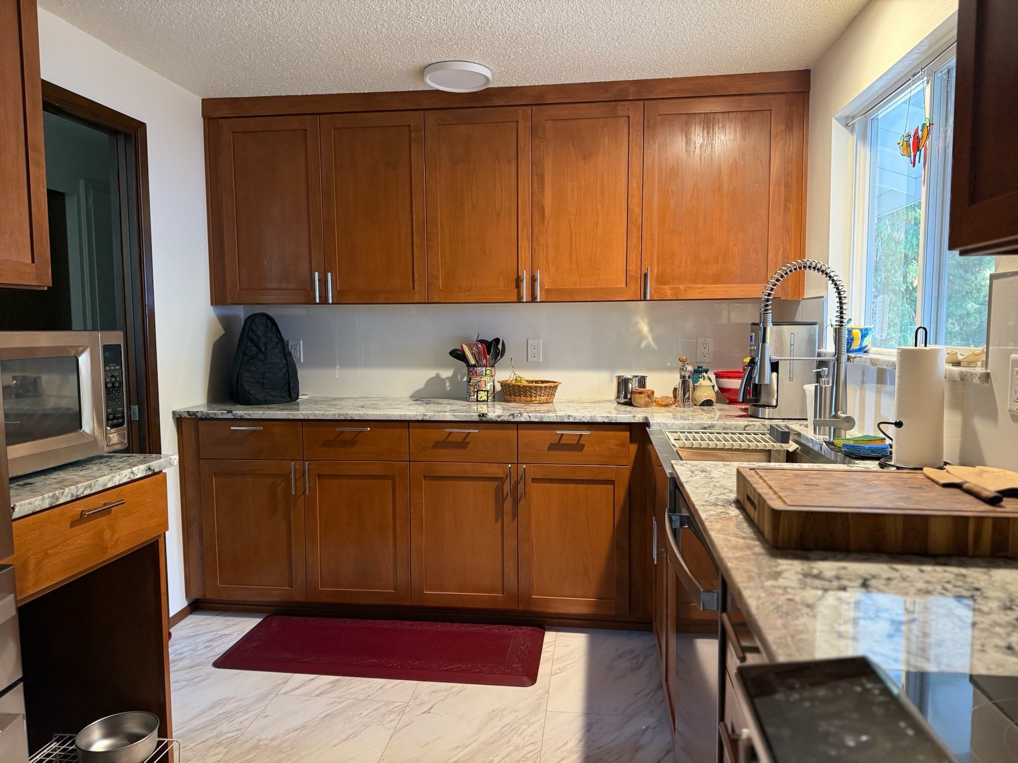 A kitchen with wooden cabinets, a marble countertop, a stainless steel microwave, and a window above the sink showing trees outside.