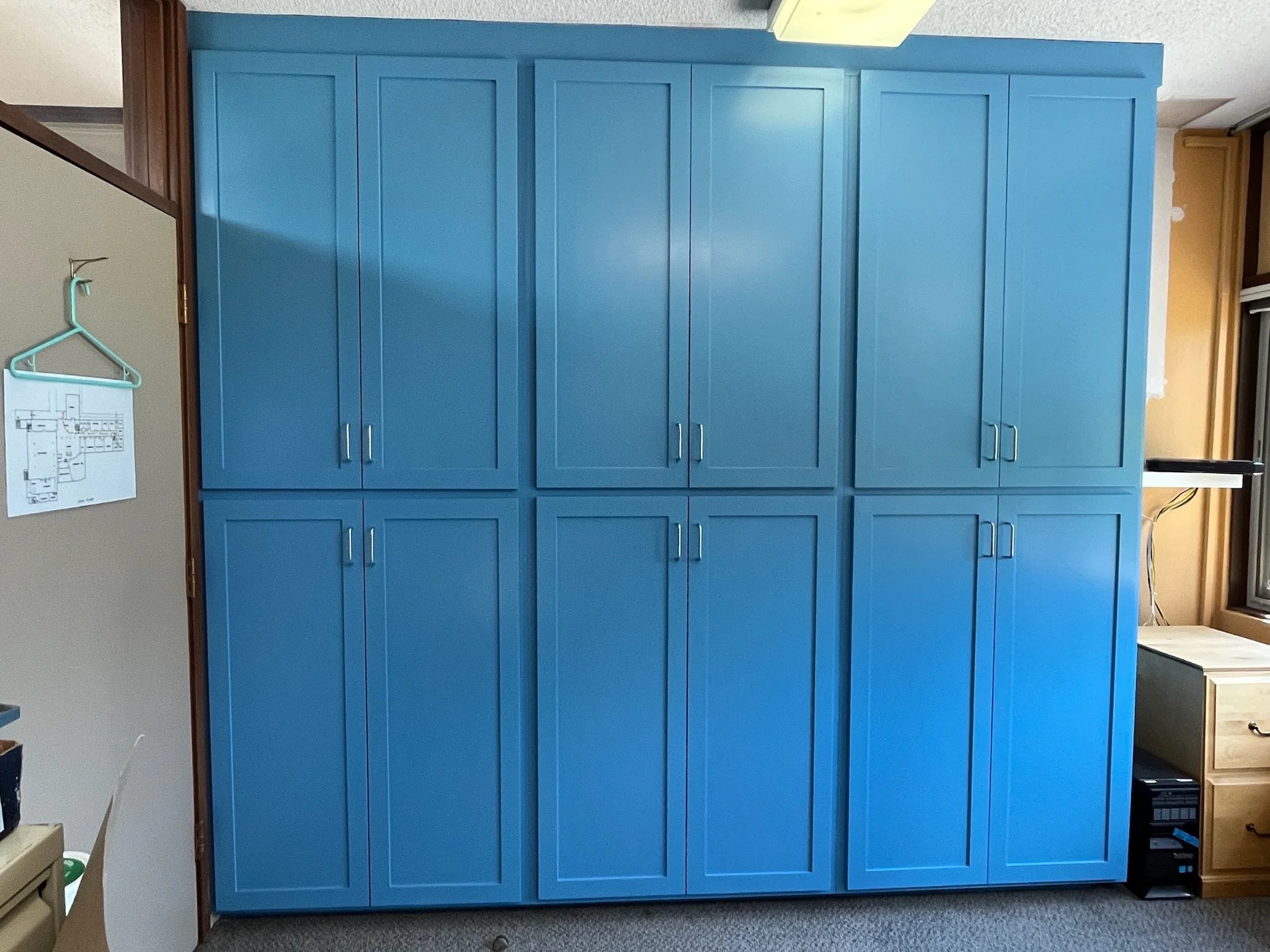 A large blue wooden cabinet with multiple doors and silver handles, set against a wall in a room with yellow and beige walls and a window.