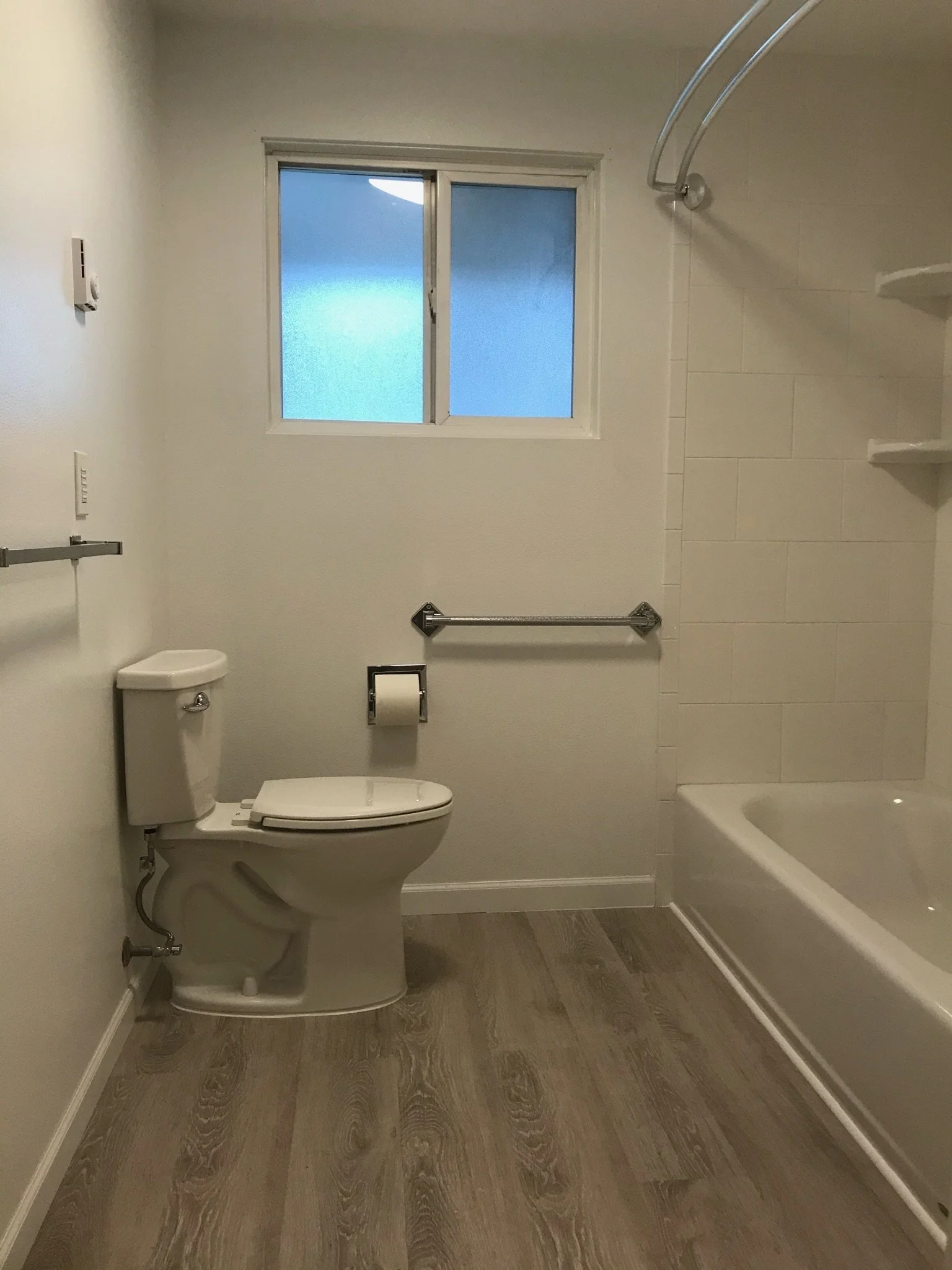 A bathroom with a window, toilet, towel bars, and a bathtub, featuring wood flooring and white walls.