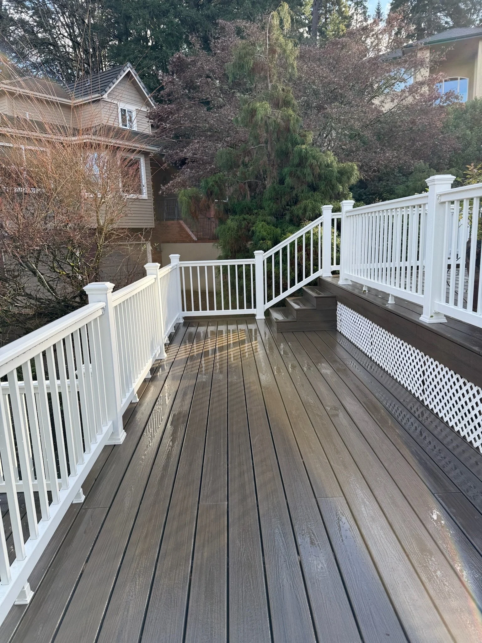 A wooden deck with white railings, stairs, and lattice skirting, overlooking neighboring houses and trees.