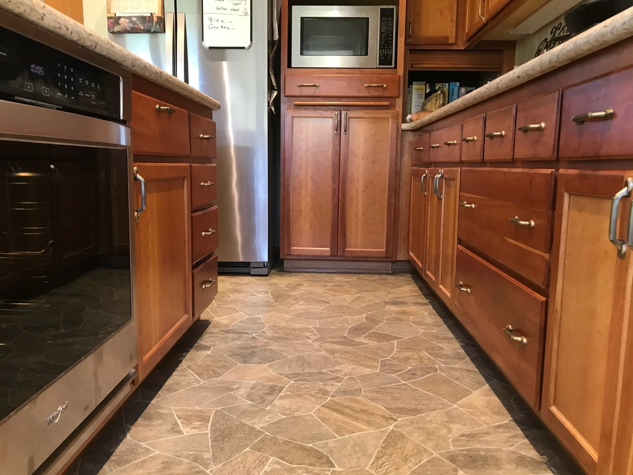 The image shows a narrow kitchen with wooden cabinets and drawers on both sides, a black stove on the left, and a stainless steel refrigerator in the background. A microwave oven is built into the cabinets above the refrigerator, and the floor has hexagonal tiles.