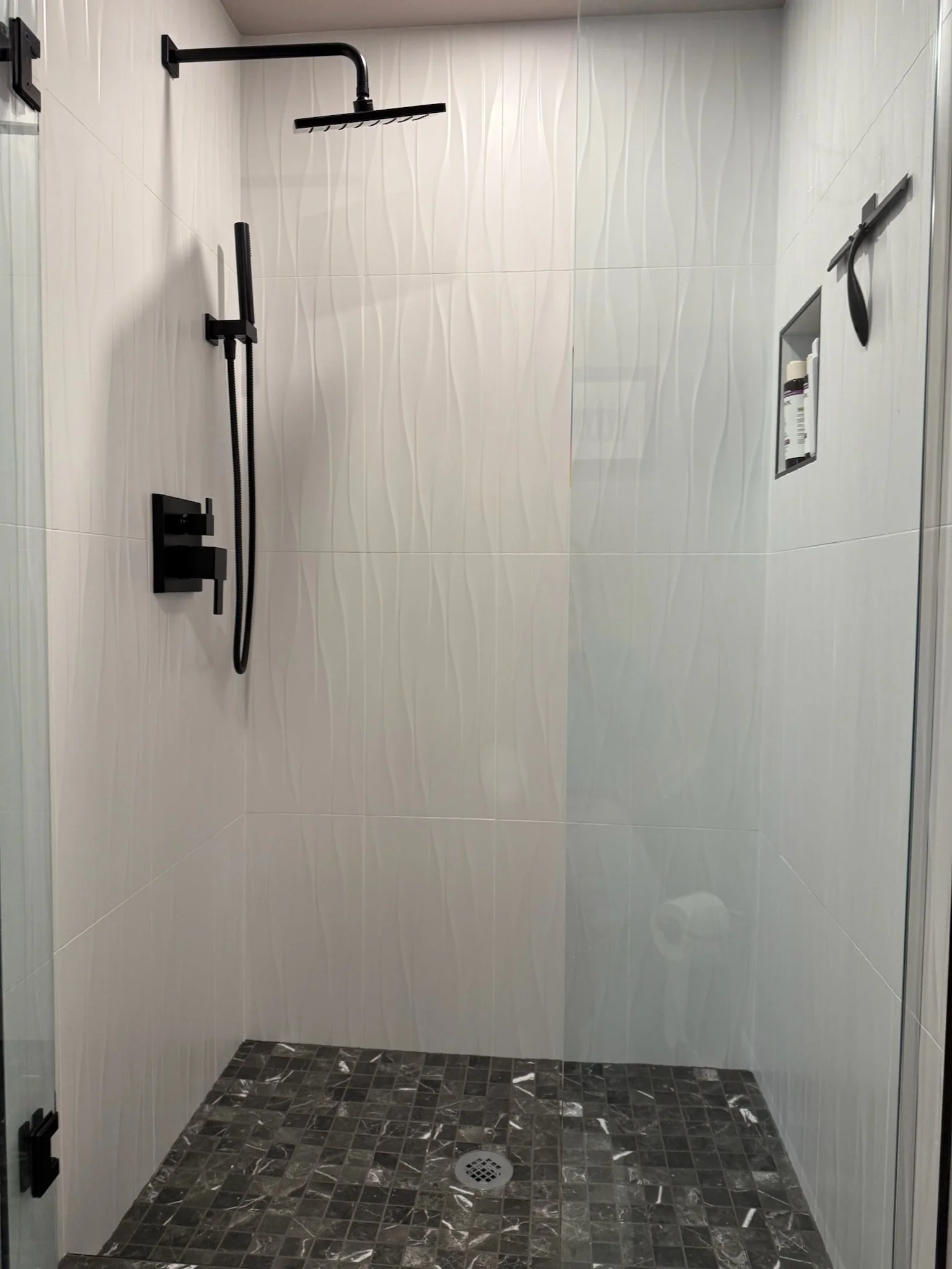 Empty modern shower with black fixtures, white tiled walls with wavy design, and a dark tile floor with a circular floor drain.
