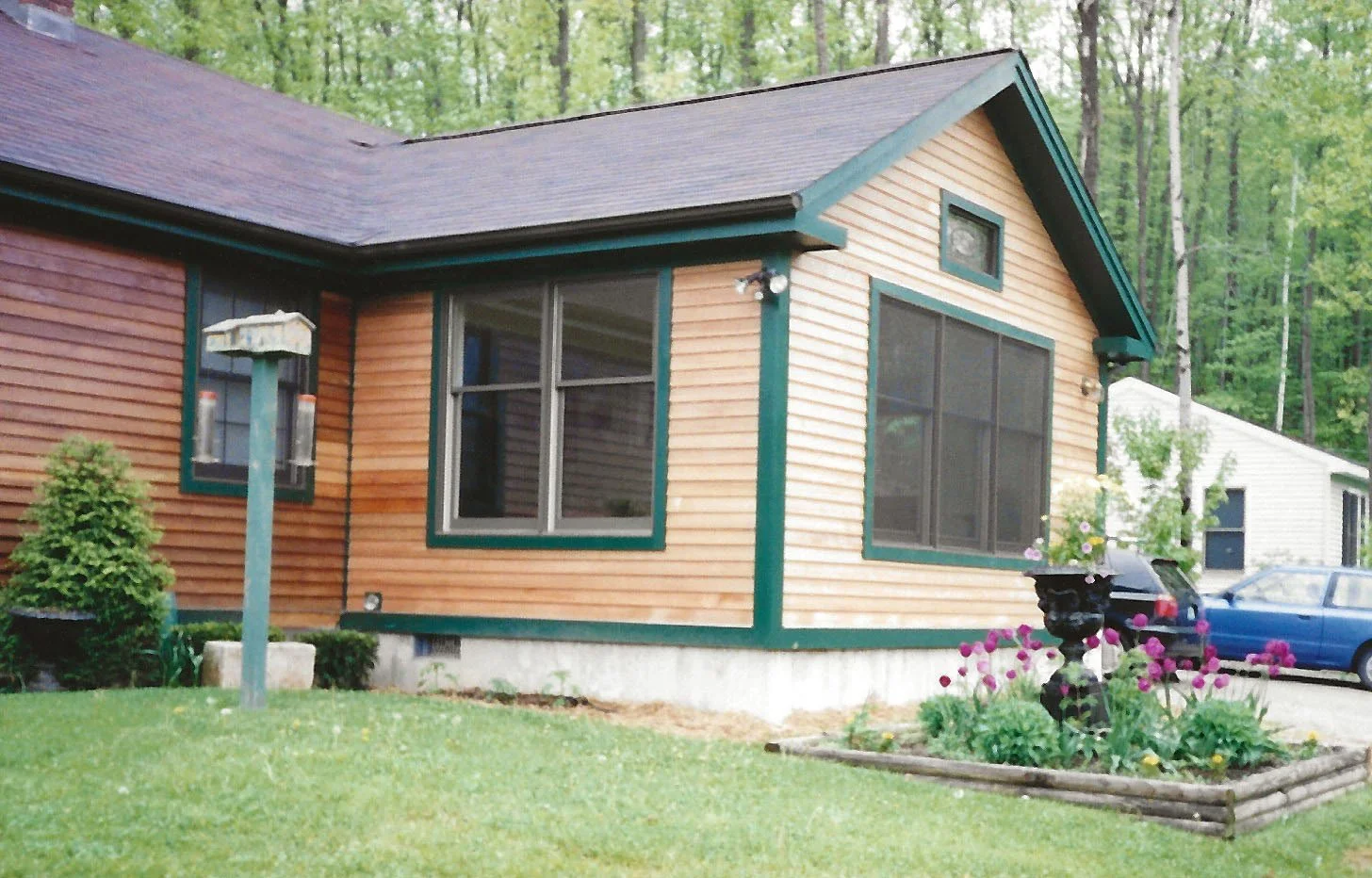 A wooden house with a sloped roof and green trim around the windows, located in a wooded area, with a manicured lawn, a black decorative planter with pink flowers, and two parked cars.