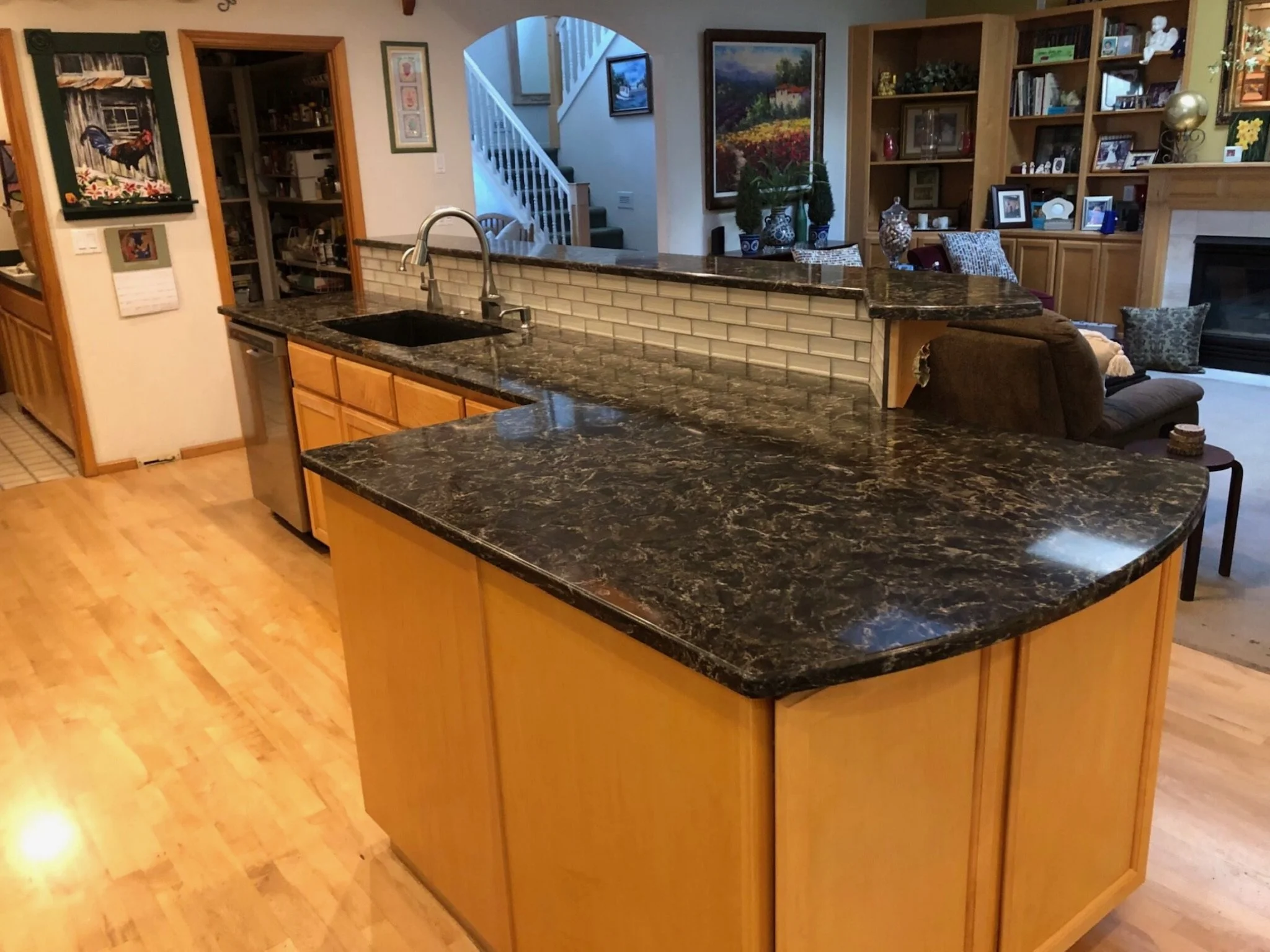 Interior view of a kitchen with a dark granite countertop, wooden cabinets, a sink with a silver faucet, and a beige brick backsplash. Adjacent to the kitchen is a living area with bookshelves, framed pictures, and decorative items, visible through an open-counter window.