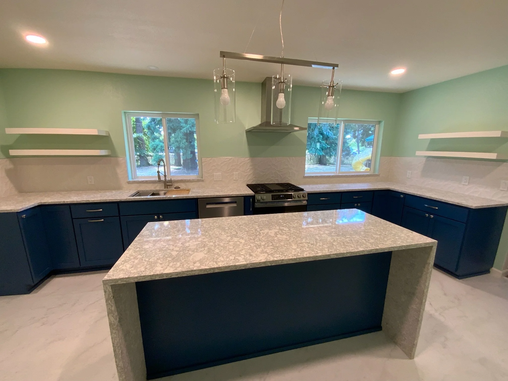 Modern kitchen with light green walls, two large windows, blue lower cabinets, white marble countertops, pendant lights, and a stove.
