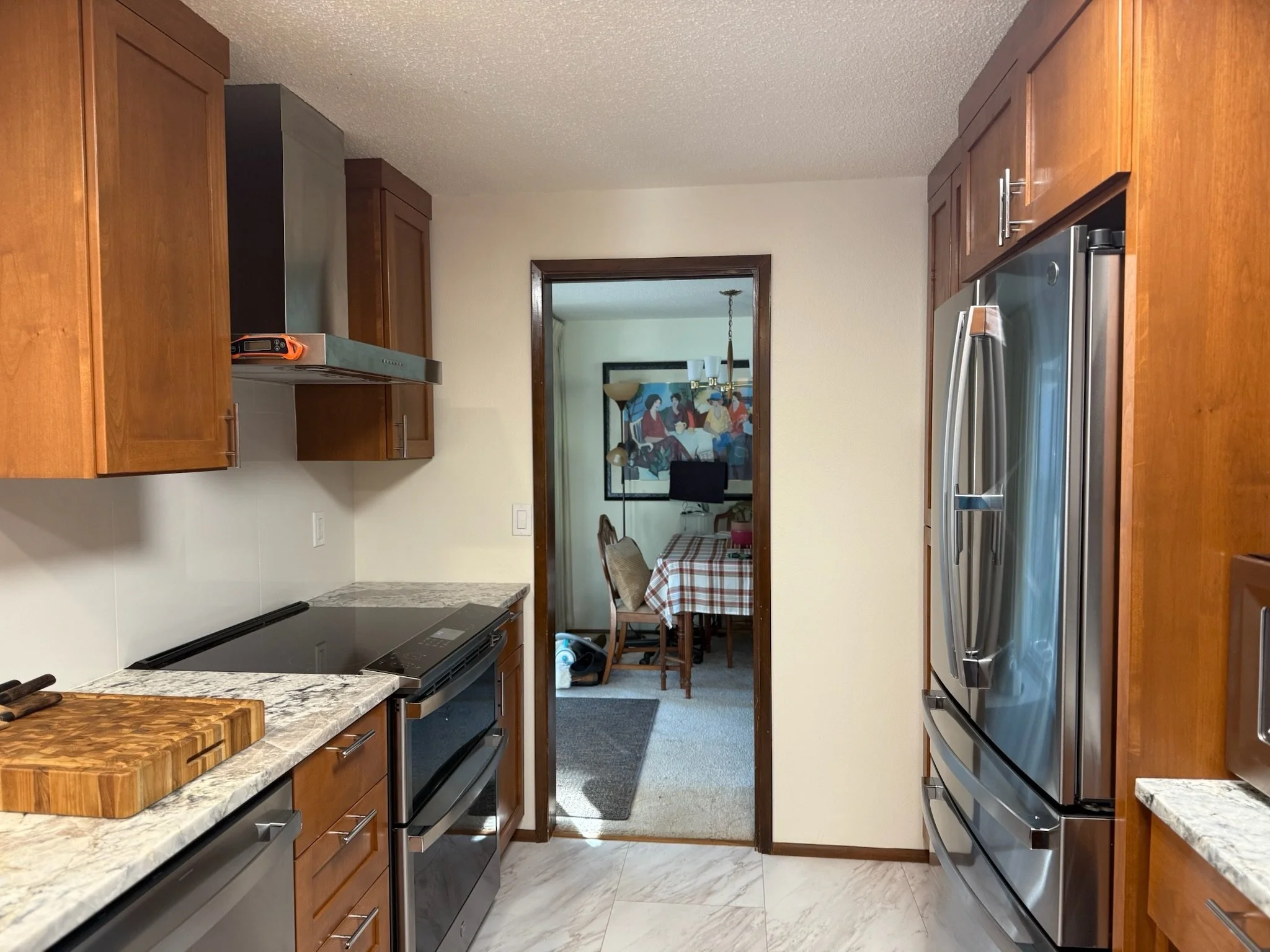 View of a kitchen with wooden cabinets, stainless steel appliances, and a marble countertop, looking into a dining area with a table and chairs.