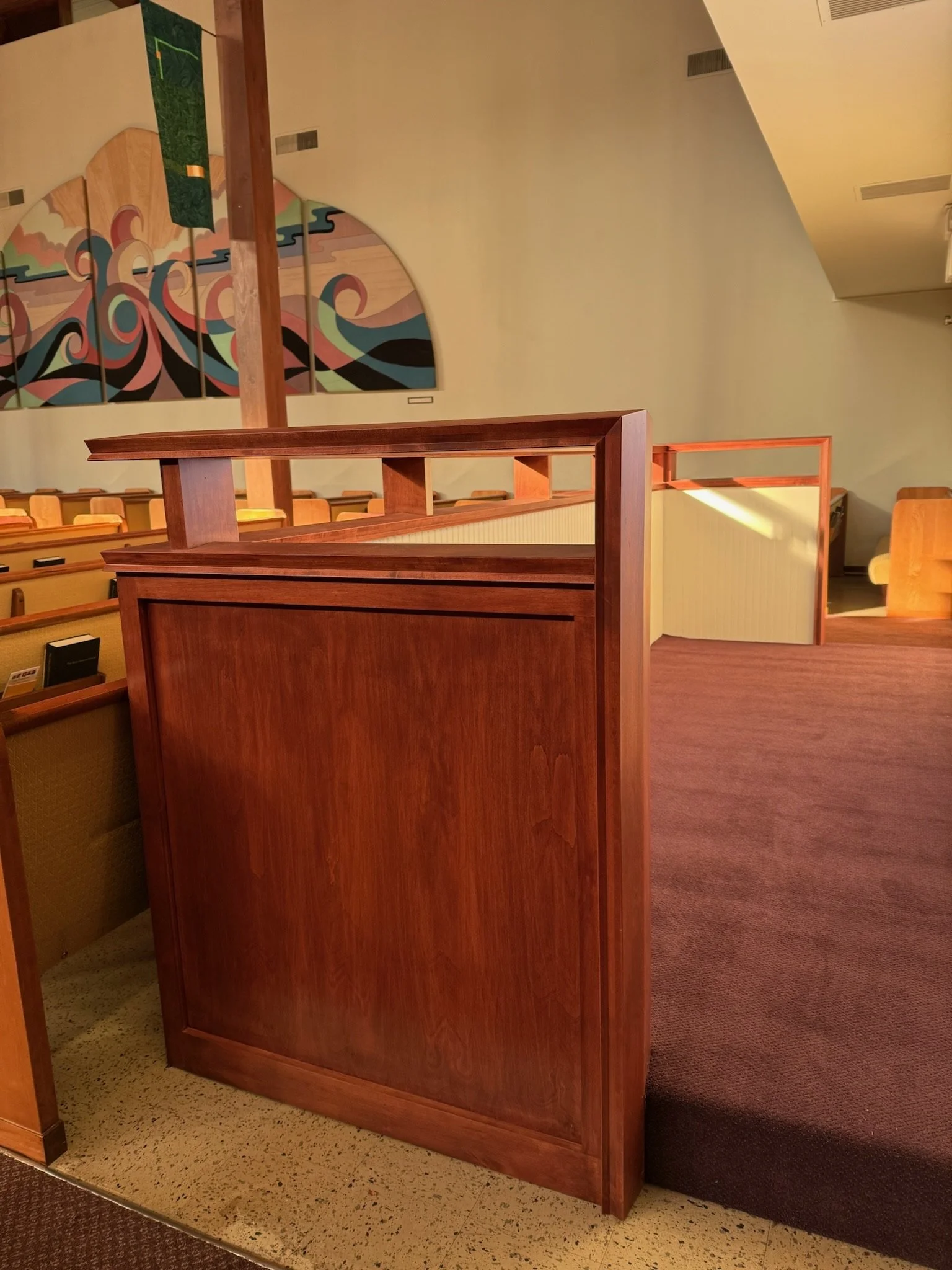 View of a church interior with a wooden pulpit, yellow upholstered pews, a colorful wall mural with swirling patterns, and a beige carpeted floor.