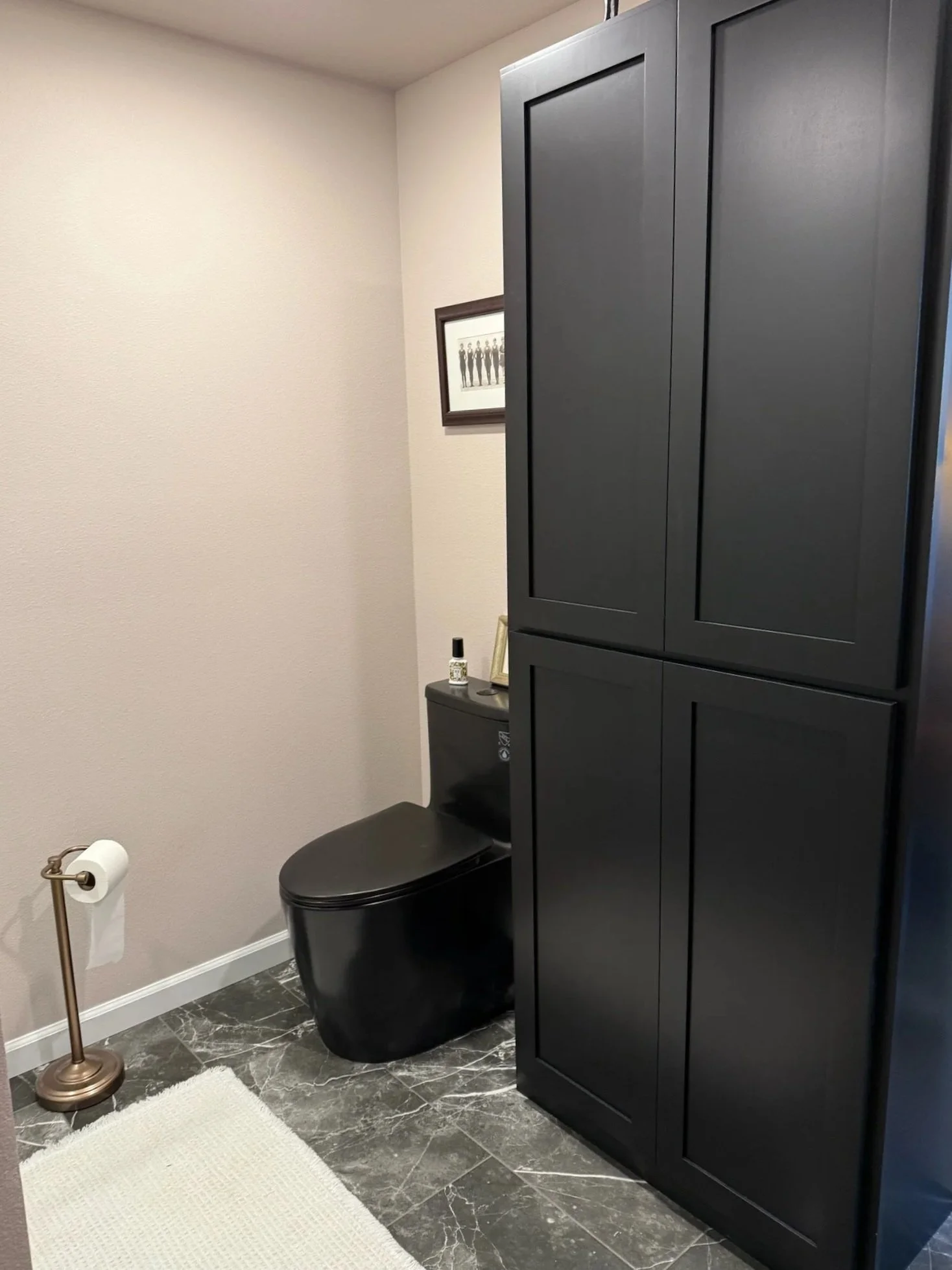 A modern bathroom with a black toilet, black cabinet, and gray marble floor. There is a white rug, a gold toilet paper holder, and a framed picture of people on the wall.