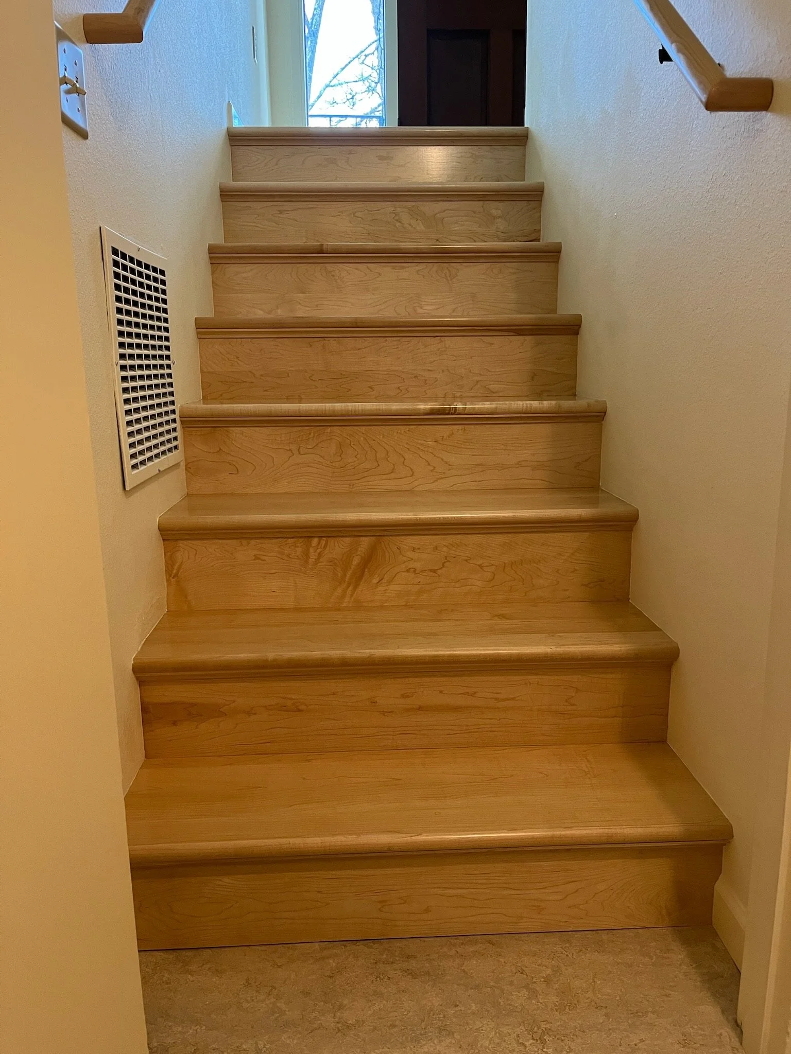 Wooden staircase with beige walls and a vent on the left side, leading to a doorway with a view of trees outside.