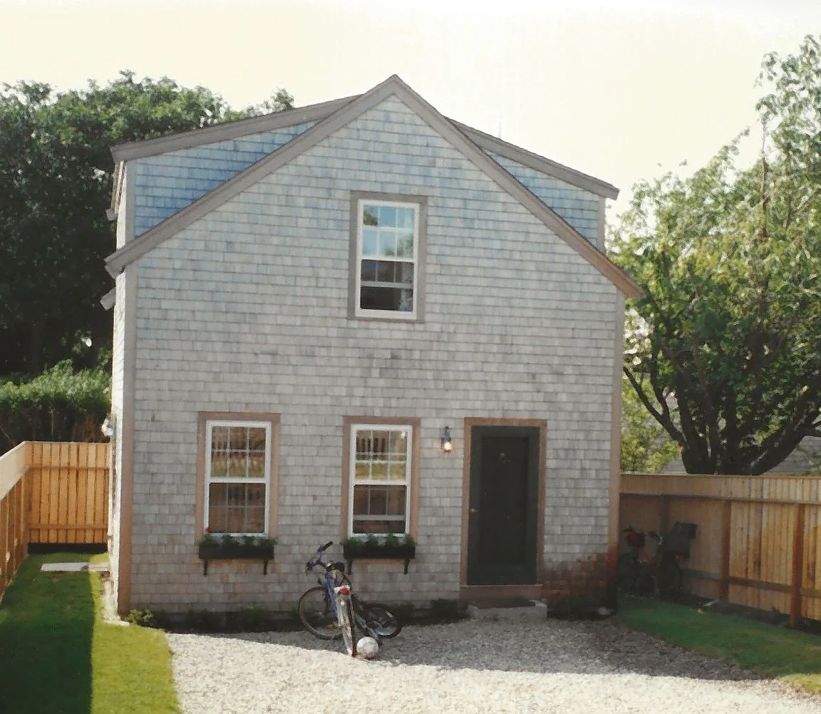 A two-story house with gray siding, three windows on the front, a black door, a bicycle parked outside, and a wooden fence on the left and right sides, with a gravel pathway and green lawn.