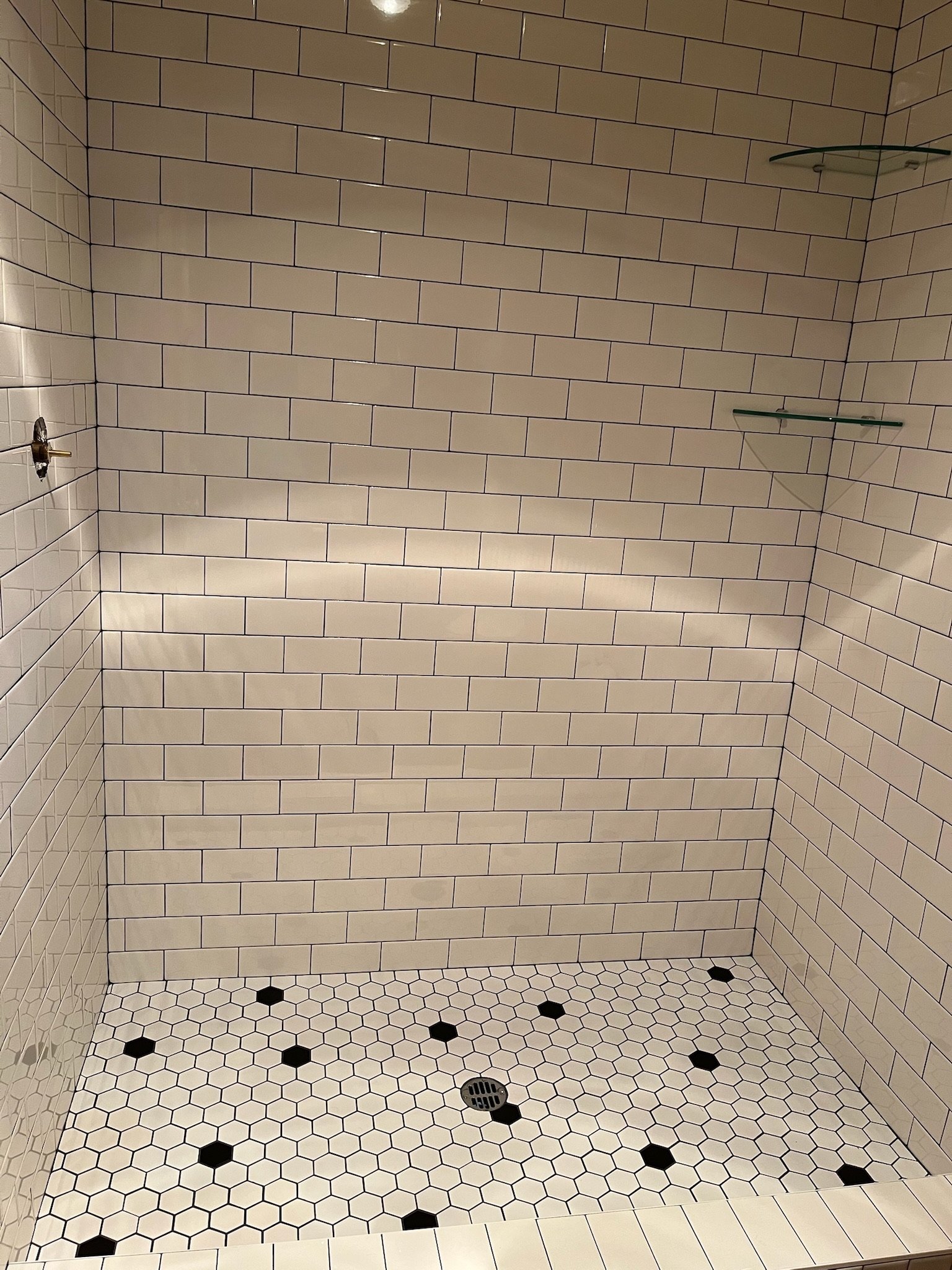 A tiled shower stall with white subway tiles on the walls, black and white hexagonal tiles on the floor, a drain in the center, and two glass corner shelves installed on the right wall.