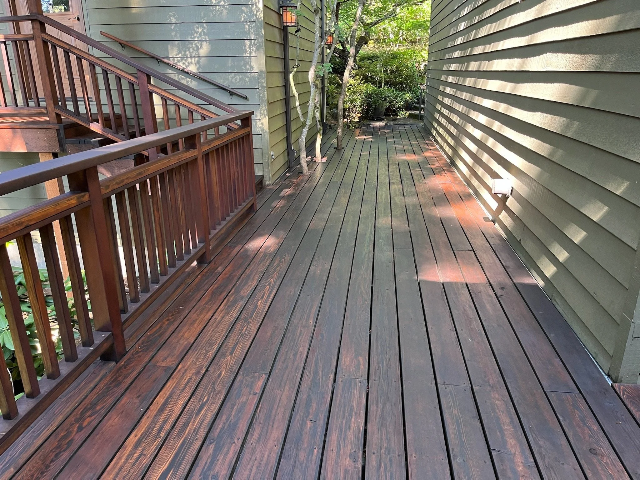 A wooden deck with a railing on the left side and trees with green foliage in the background. The siding of the house is painted green with horizontal lines, and there are outdoor lights on the wall.