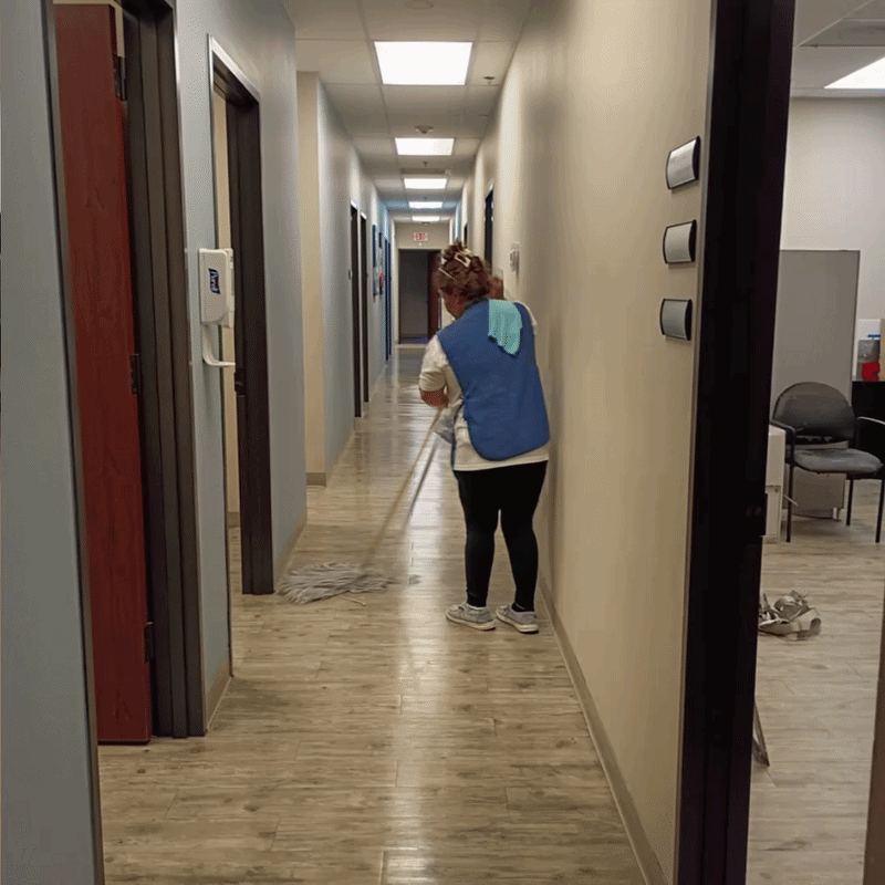 A woman cleaning a long, narrow office hallway with a mop, wearing a blue vest and black pants.