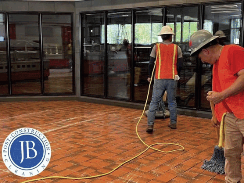 Construction workers cleaning a tiled floor inside a building with glass walls and doors. One worker is using a mop, and another is holding a power washer, with a yellow extension cord on the floor.