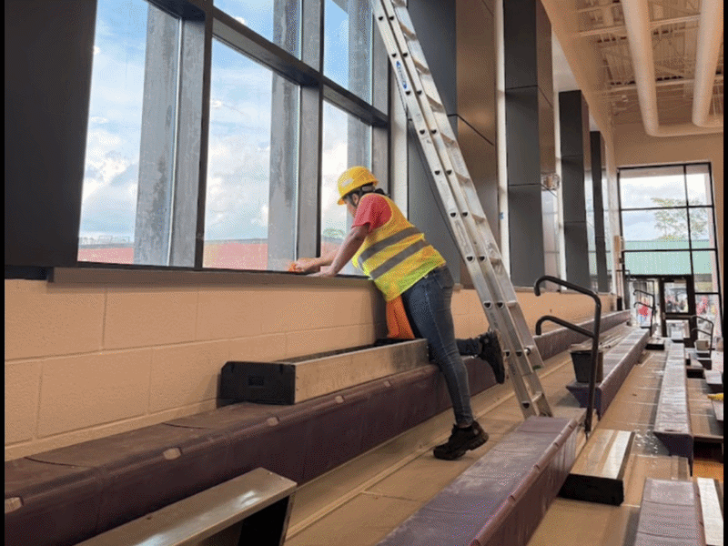 Construction worker in a yellow safety helmet and reflective vest cleaning a window in a sports arena or gym.