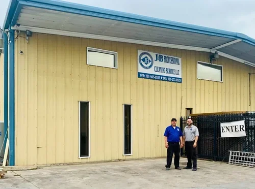 Two men standing in front of a beige industrial building with a sign that reads "JB Professional Cleaning Service LLC" and an entrance gate with a sign that says "ENTER".