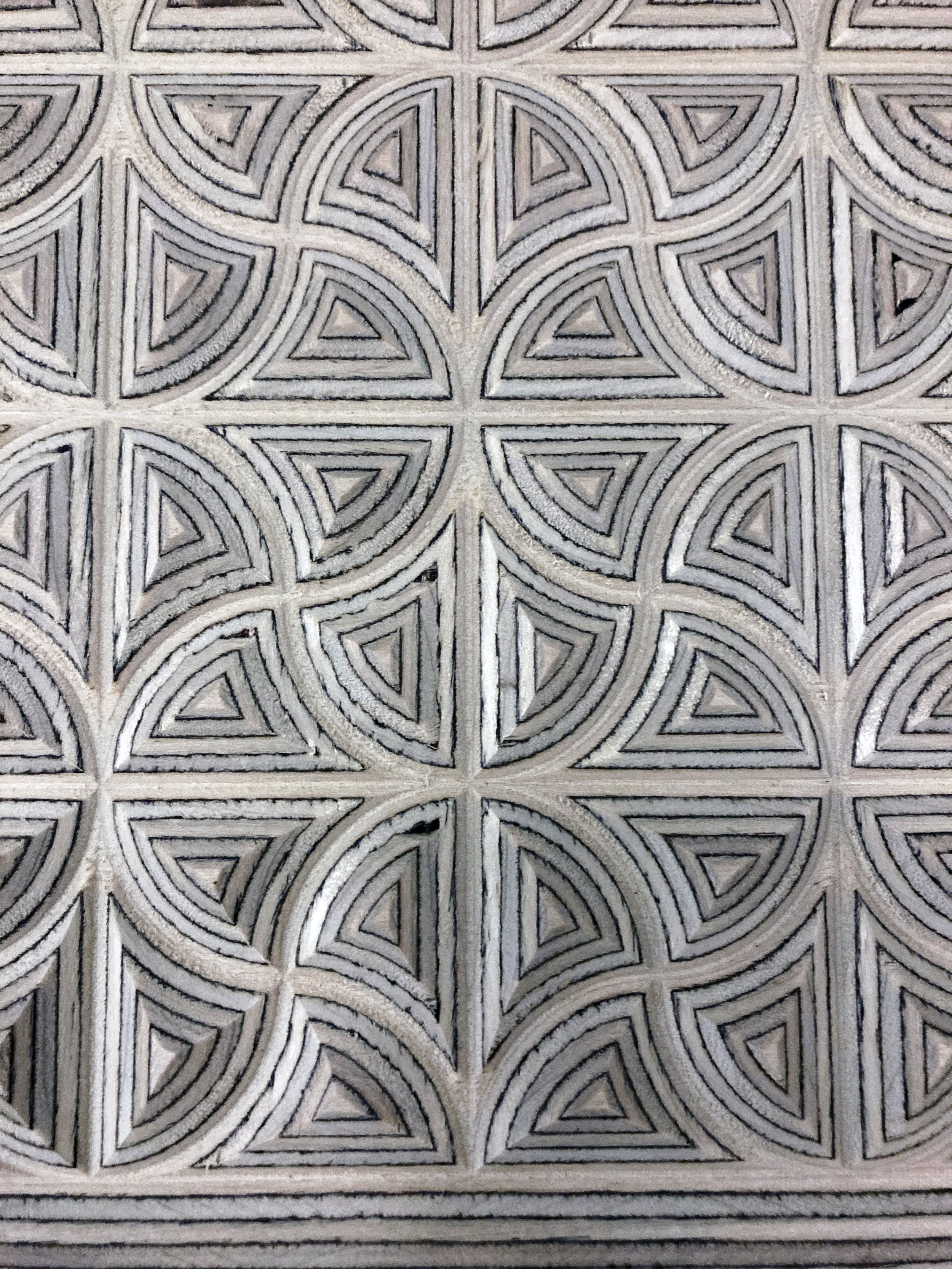 A close-up view of a detailed pattern made of carved or embossed wood, featuring repeating geometric shapes with curved lines and triangular motifs.