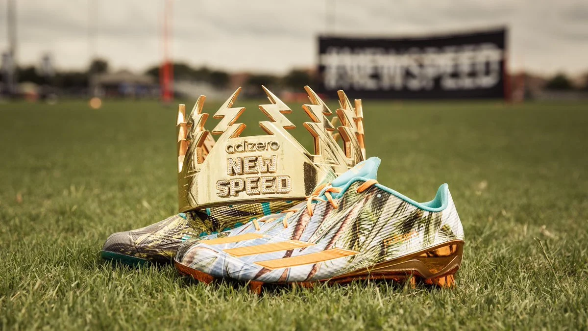 A pair of colorful soccer cleats and a gold 3d printed and painted crown set on a grassy field with a scoreboard in the background.