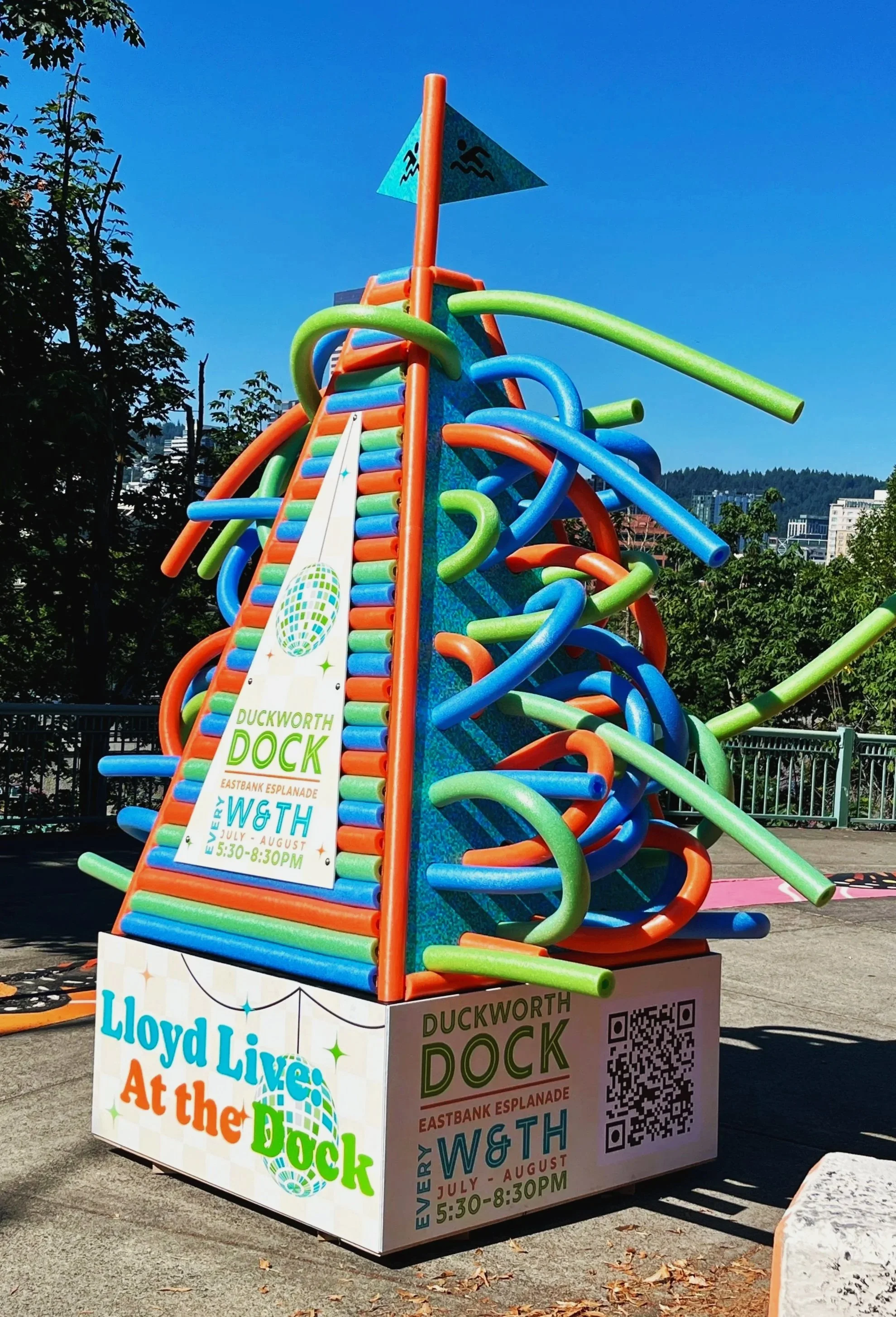 Colorful abstract outdoor sculpture made of intertwined pool noodles in orange, green, blue, and red, with a sign promoting the Lloyd Livingston event at the Duckworth Dock. Custom fabrication by MC Laser Labs for the Eco Lloyd group in Portland, OR.