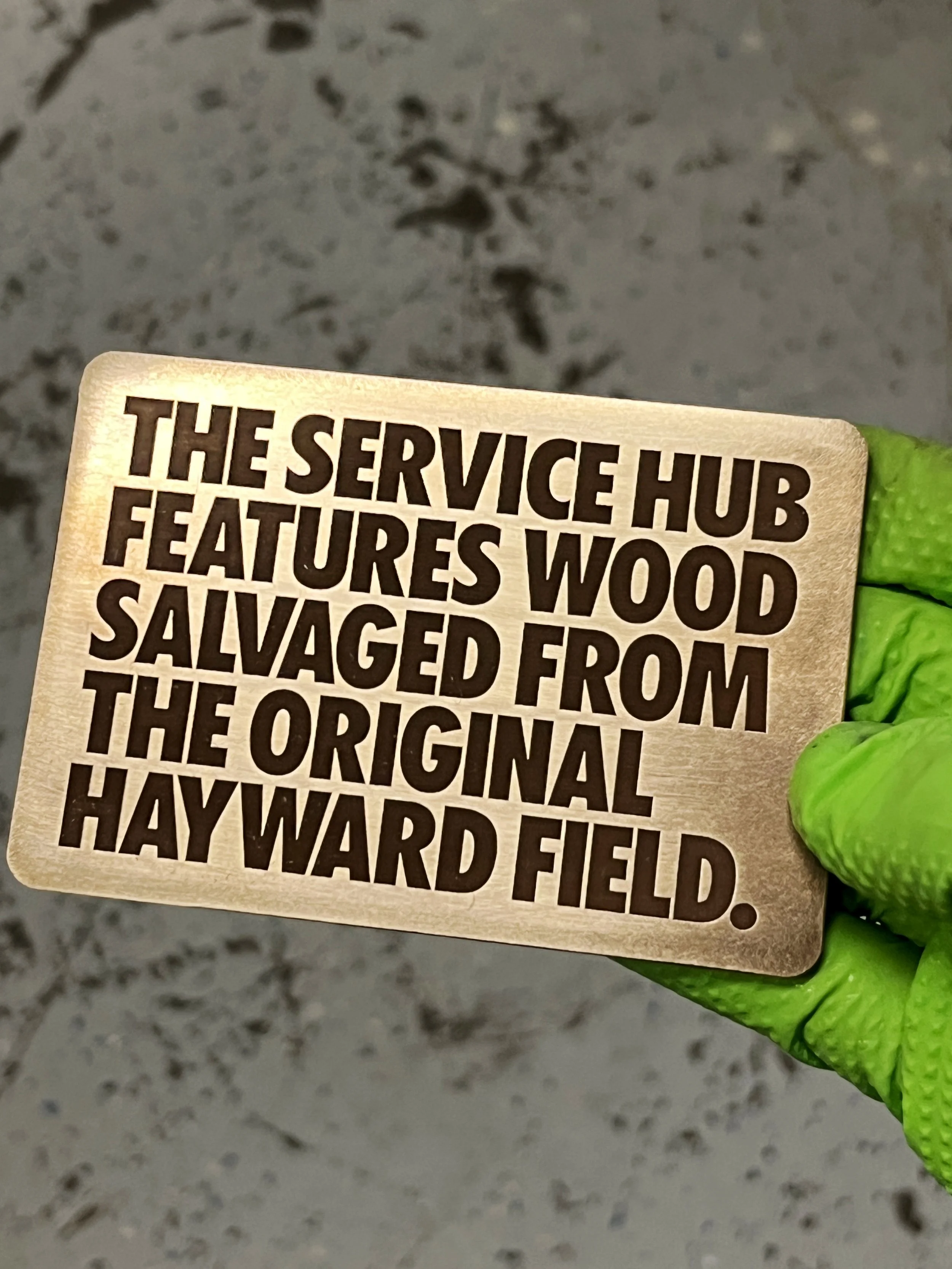 Close-up of a copper plaque for Hayward Field with laser engrvaed text.The background shows a gray, speckled surface.