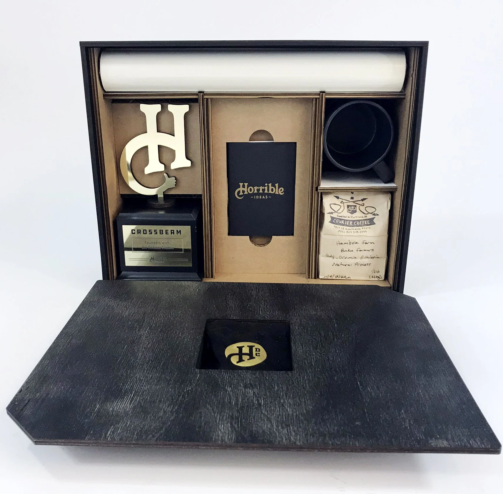 Horrible Design Co client seeding kit box, black reception desk display with woodgrain texture, napkin holder, 'Horrible Ideas' book, mug, and gold 'H' and '$' letters, custom fabrication by MC Laser Labs.