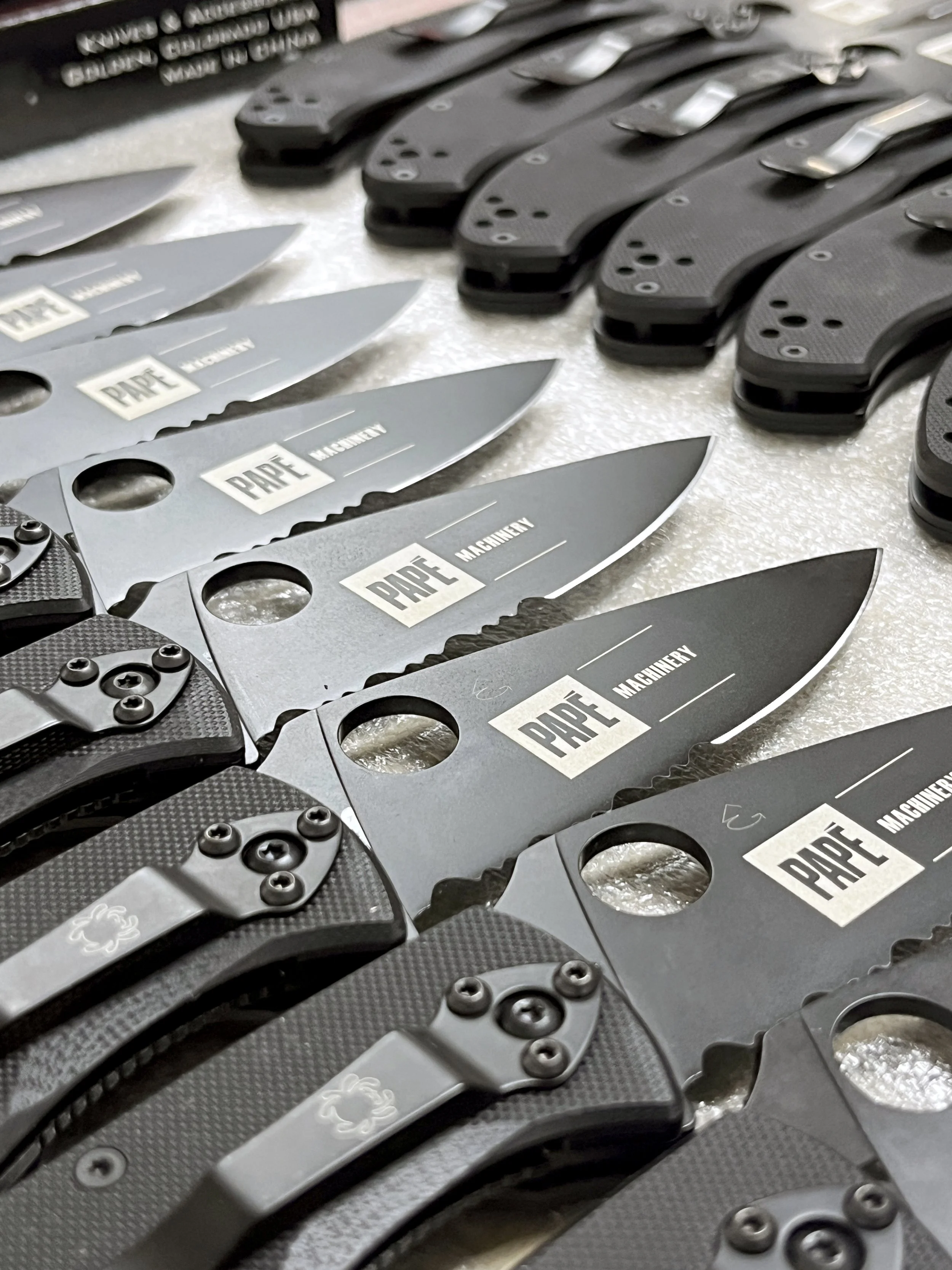 Multiple folding knives with black and gray handles, lined up on a surface. The blades are mostly gray with the word "PAPE" and "MACHINERY" written on them.