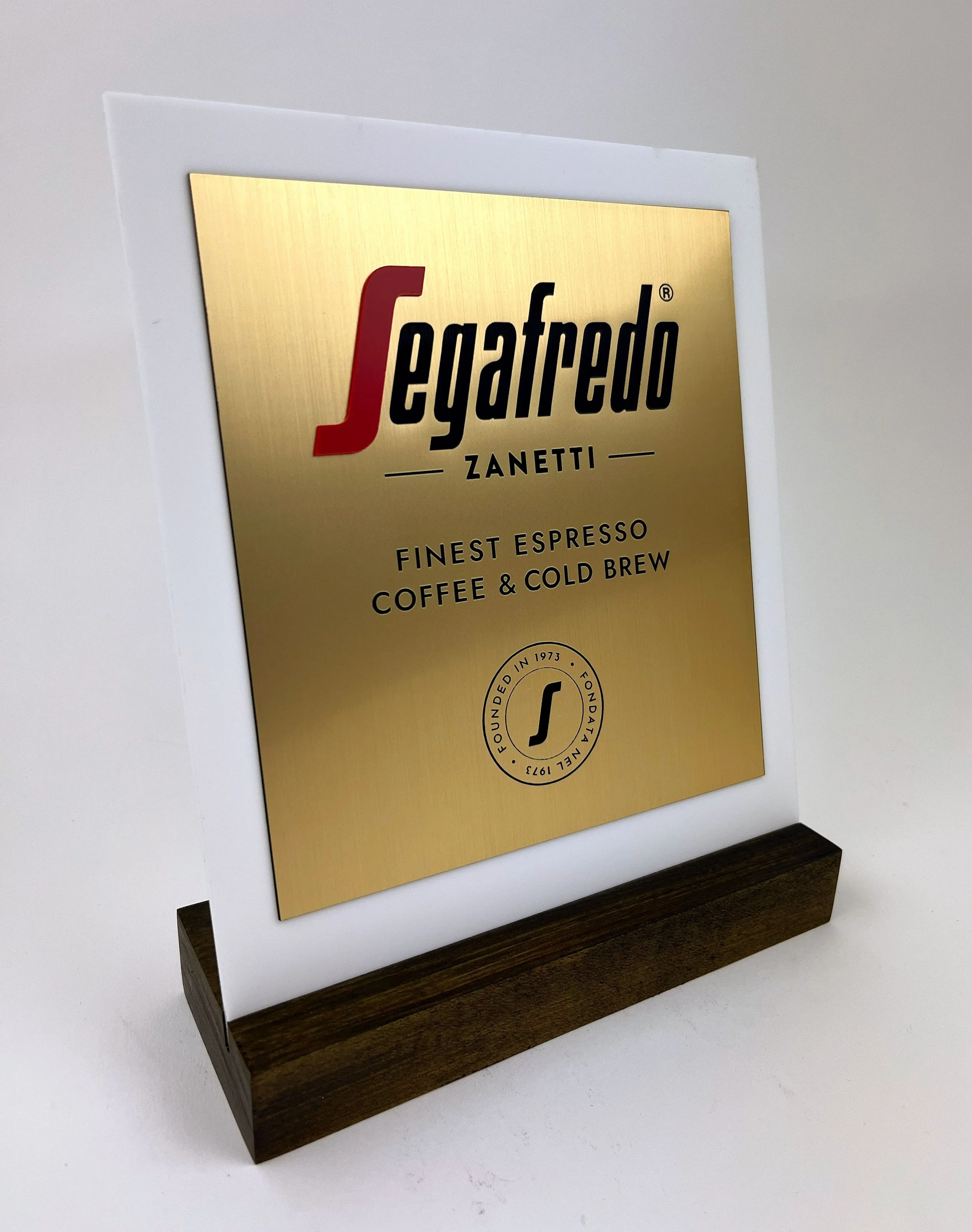 CO2 laser cut and engraved gold-colored sign plaque for Legarego Zanetti with coffee slogan and circular emblem, mounted on a wooden stand, custom laser cutting and engraving by MC Laser Labs.