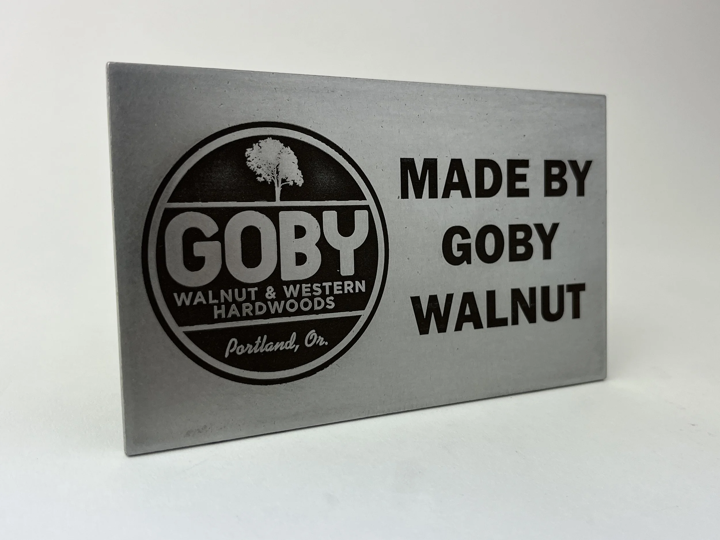 A metallic label or sign with the GOBY logo, which has a tree inside a circle, and the words "WALNUT & WESTERN HARDWOODS" along with "Portland, Or." It also includes the text "MADE BY GOBY GOBY WALNUT".