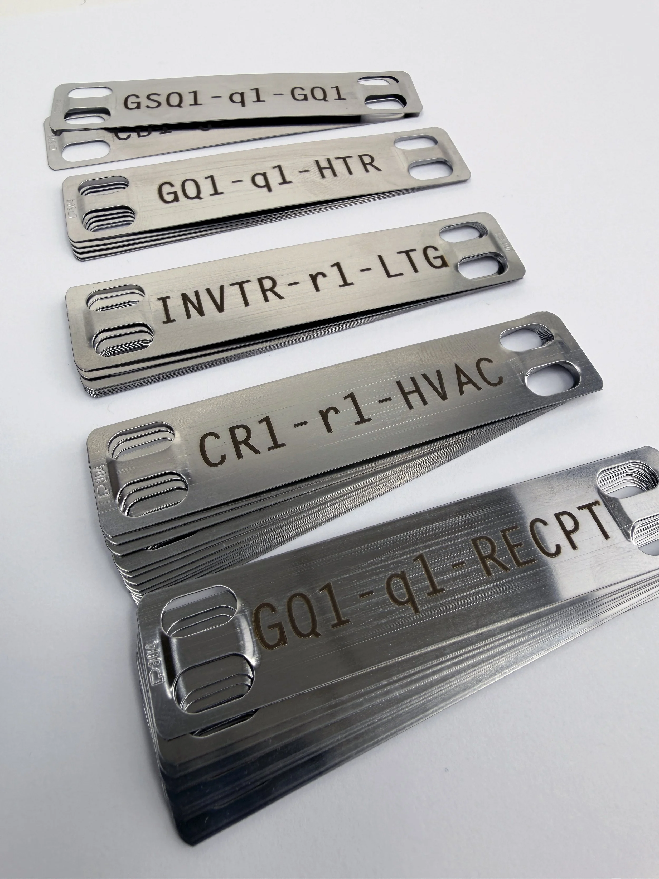 Stacked metal tags with engraved alphanumeric codes and labels such as GQ1-q1-GQ1, GQ1-q1-HTR, INVTR-r1-LTG, CR1-r1-HVAC, and GQ1-RQ1-RECPT, each with two oval holes on the left side.