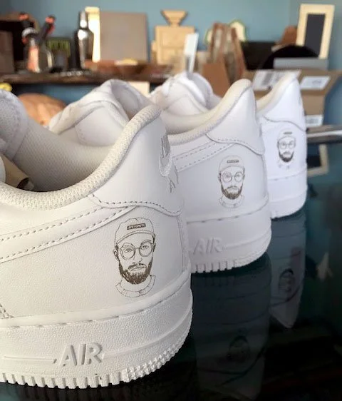 Custom printed white sneakers with a man's face line drawing on the heel, lined up in a store display, custom printing by MC Laser Labs.