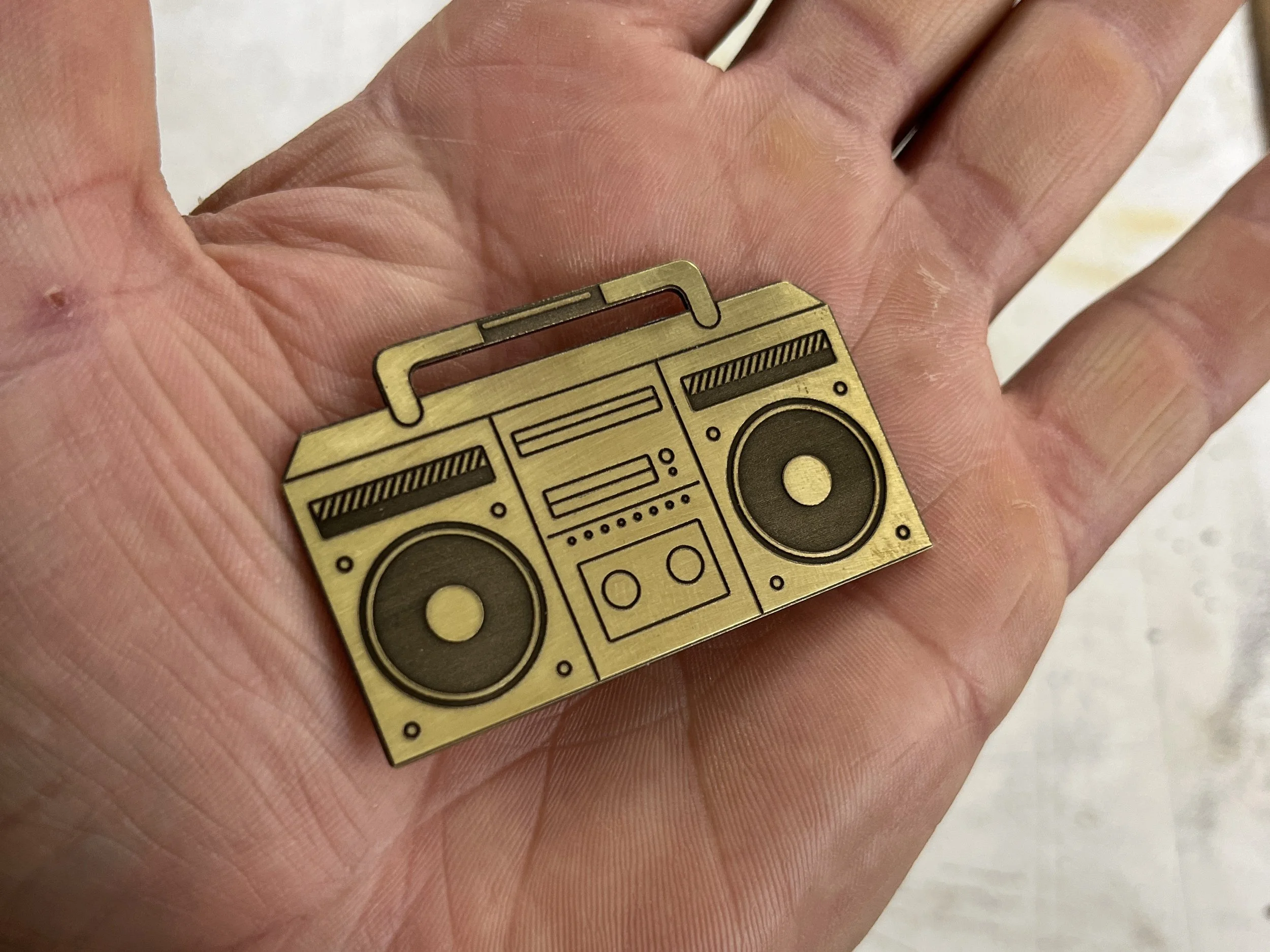 A close-up of a person's hand holding a gold-colored rectangular badge with black printed design features of a stereo system, including speakers, a control panel, and buttons.