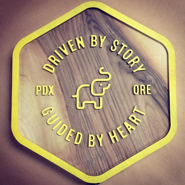 Decorative sign with yellow hexagonal border, featuring a small elephant illustration in the center, and the text 'Driven by Story, Guided by Heart' and 'PDX' and 'ORE' around the elephant.