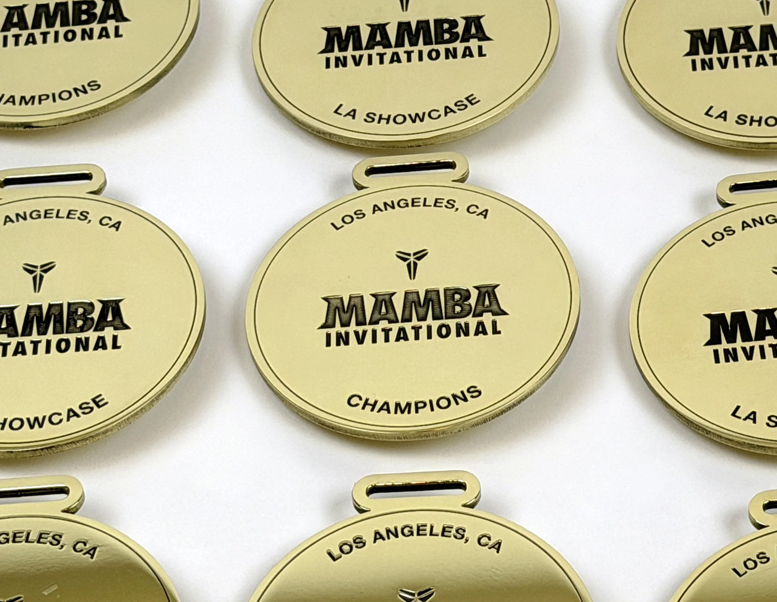 Close-up of several laser engraved brass medals with black text that reads "Mamba Invitational, La Showcase, Champions, Los Angeles, CA," and a black logo, arranged on a white background.