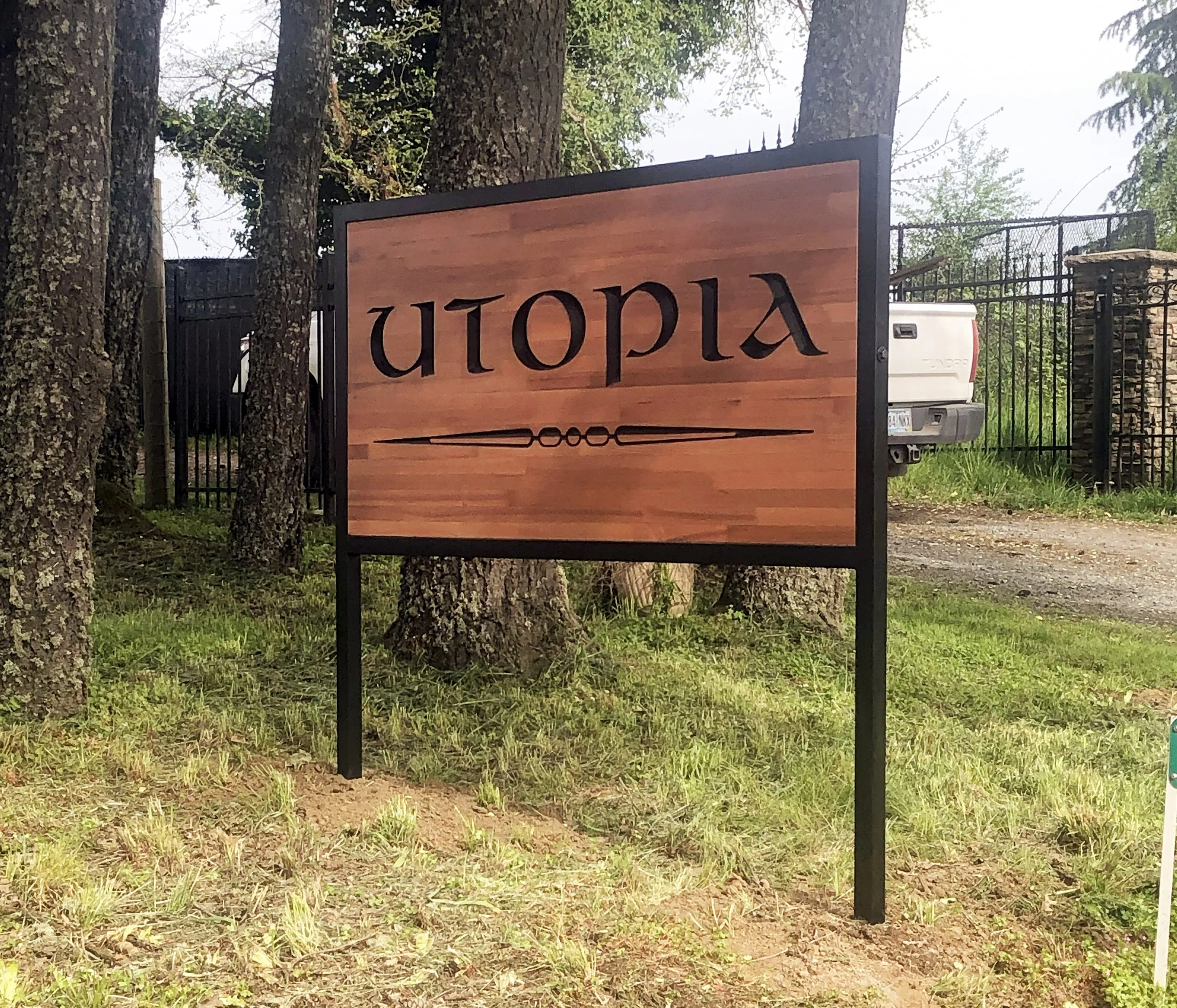 Wooden sign with black letters and arrow, mounted on black metal frame, surrounded by trees and grass.