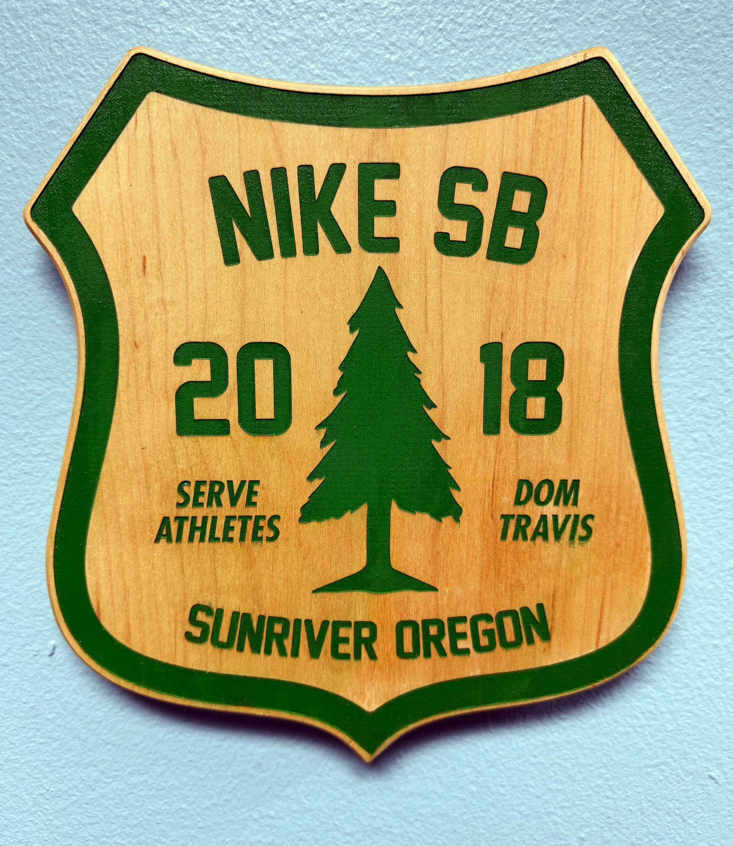 Wooden sign with green border and text, featuring a pine tree illustration, indicating Sunriver, Oregon, serving athletes, and encouraging outdoor activities.