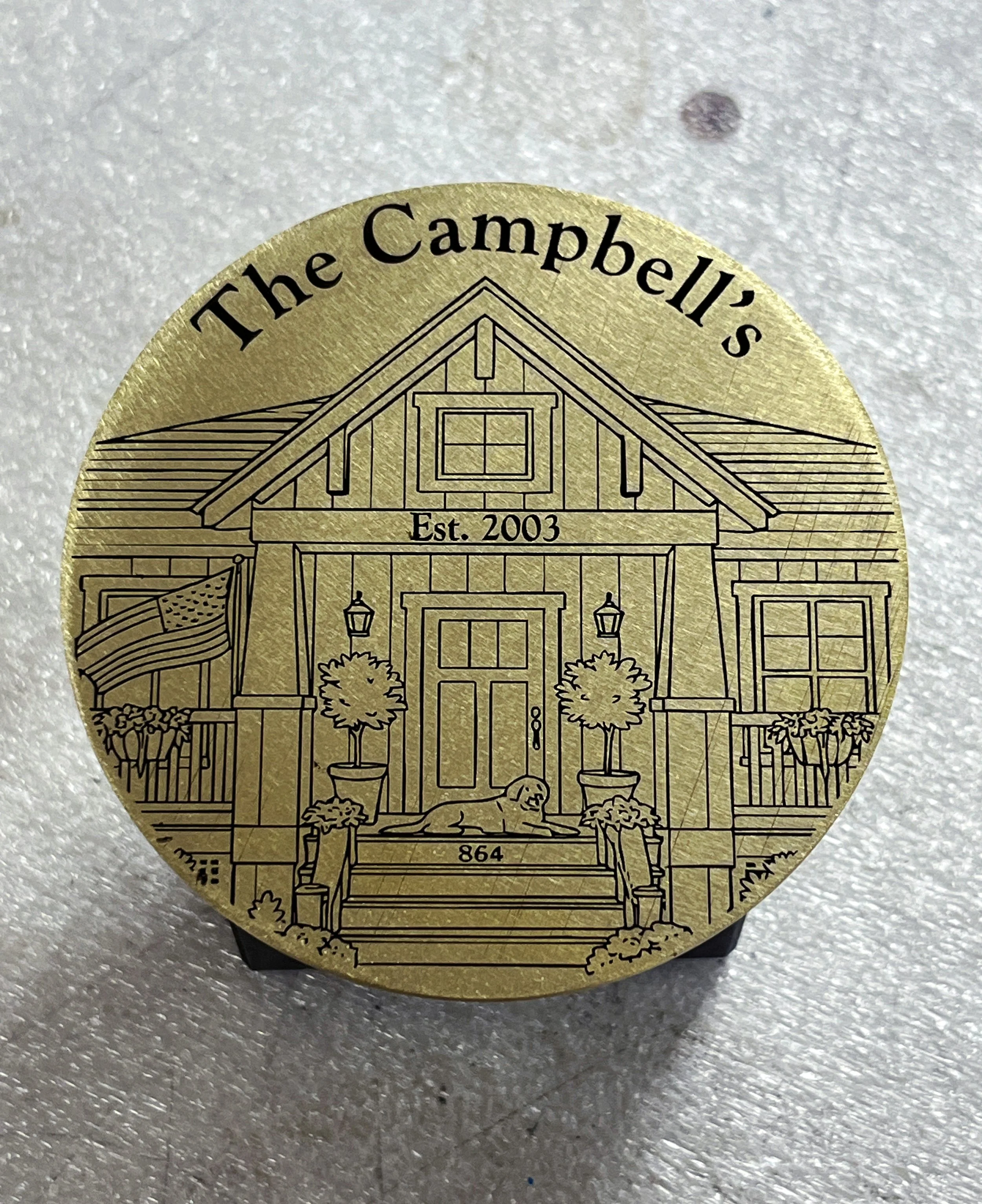 A gold-colored round ornament featuring a detailed line drawing of a house with a peaked roof, front porch, and potted plants, with the text "The Campbell's" at the top, "Est. 2003" in the middle, and the number "864" at the bottom.