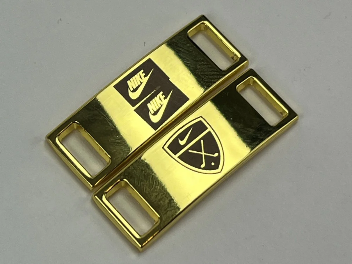 Two gold-colored metal plate clips with logos, one with Nike logo and the other with Nike shield logo.