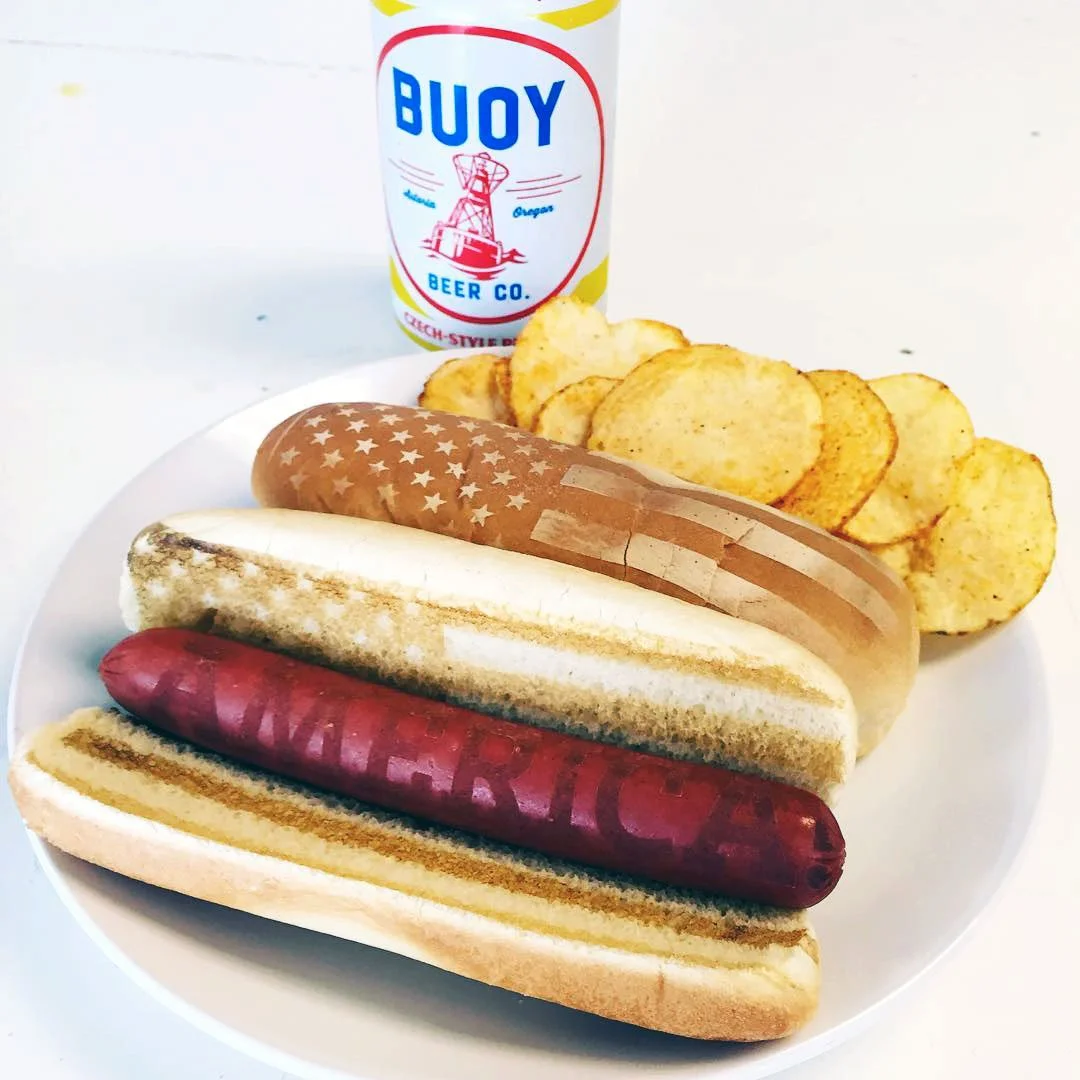 Plate with three hot dogs in buns, potato chips, and a can of Buoy Beer.