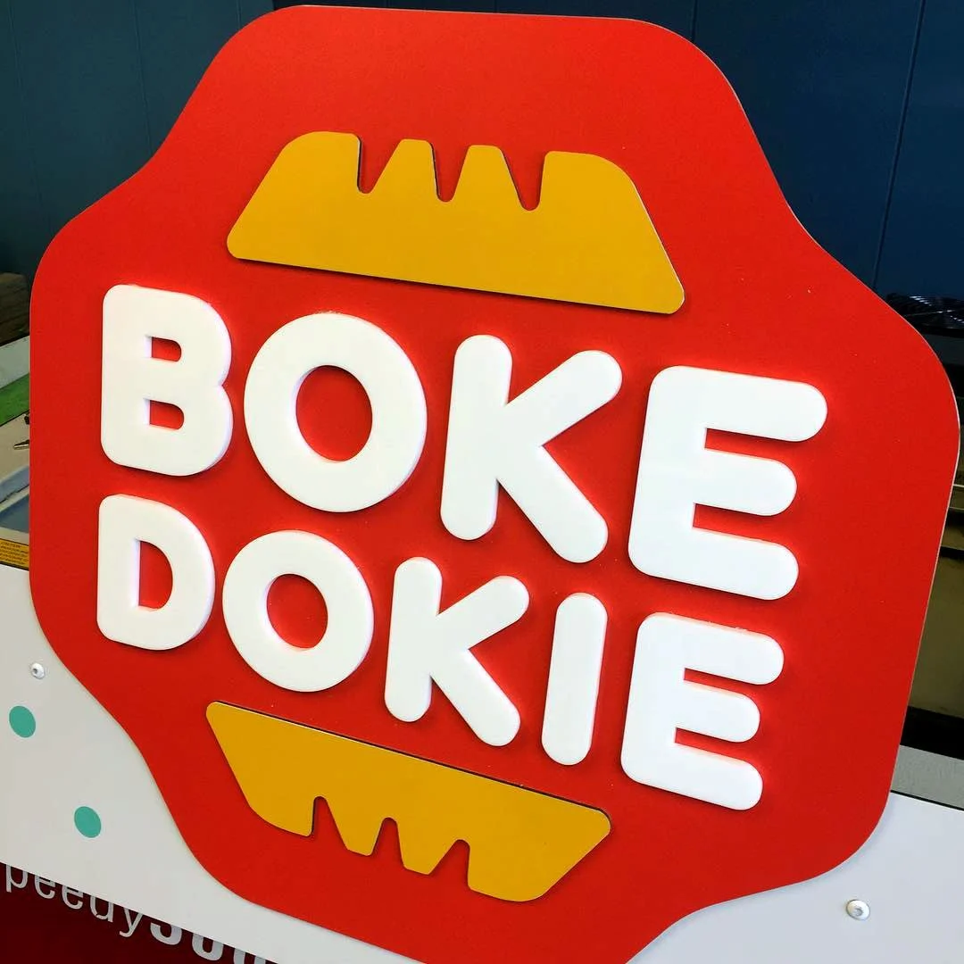 CO2 laser cut and engraved red sign with white 3D letters reading 'BOKE DOKIE' and yellow waffle icons, custom laser cutting and engraving by MC Laser Labs.