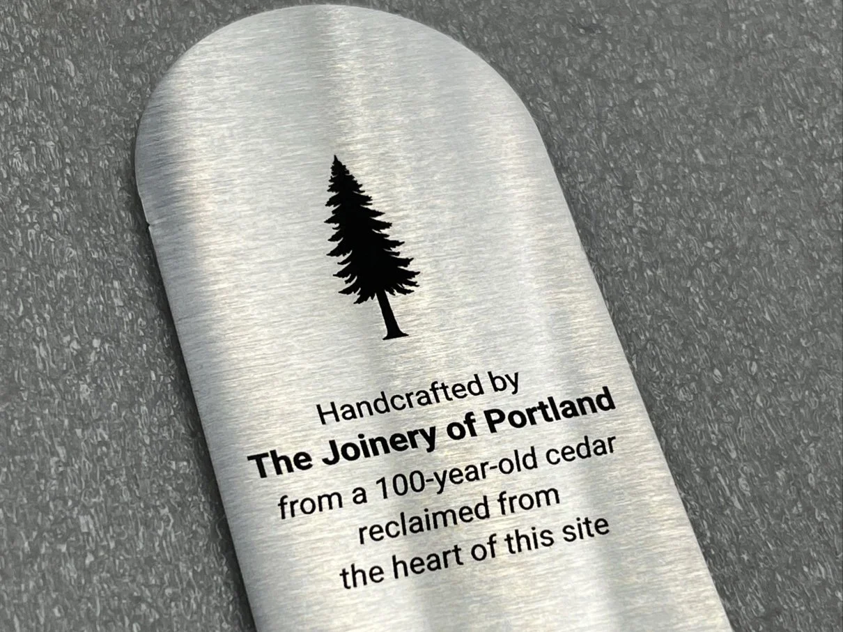 A metal product with a black pine tree illustration at the top. Text reads: 'Handcrafted by The Joinery of Portland from a 100-year-old cedar reclaimed from the heart of this site.'