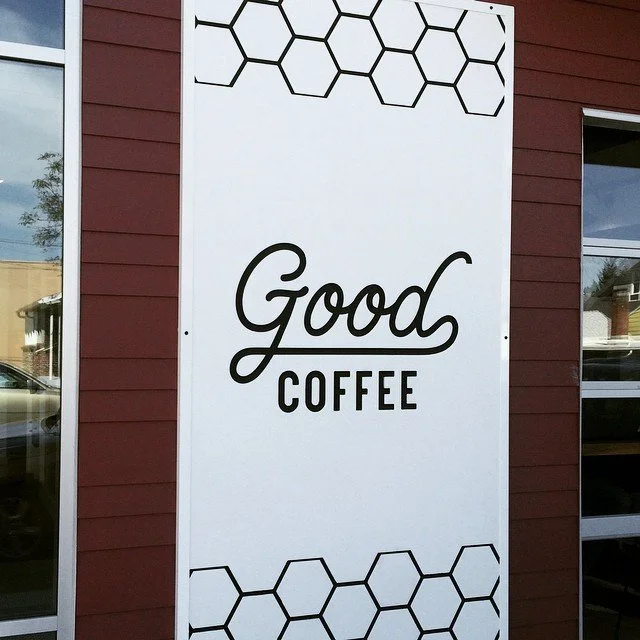 Sign reading 'Good Coffee' with hexagon patterns at the top and bottom, mounted on a red wall outside a building.