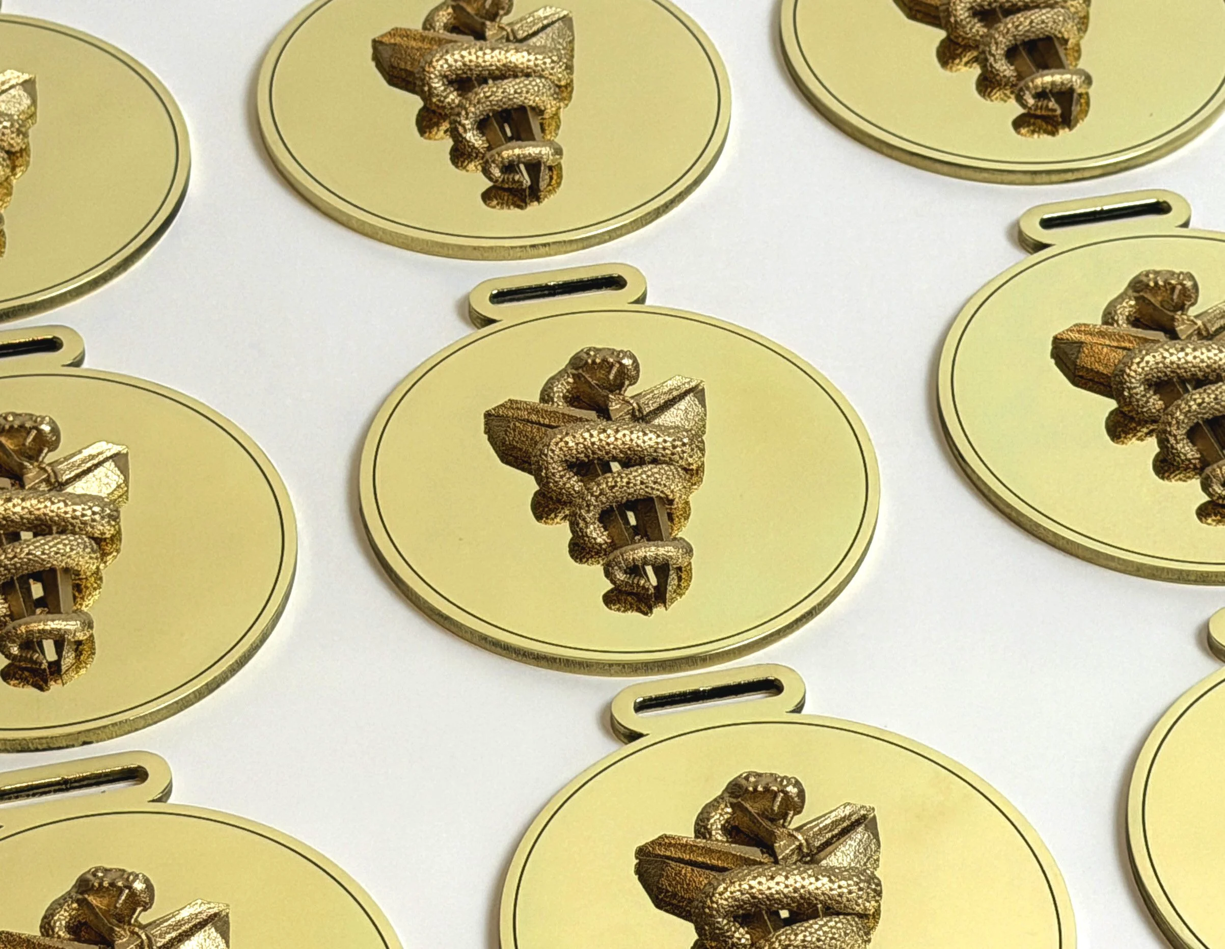 Close-up of several brass Mamba League Invitational medals with a snake wrapped around a chalice design that is 3d Printed on each, arranged on a white surface.